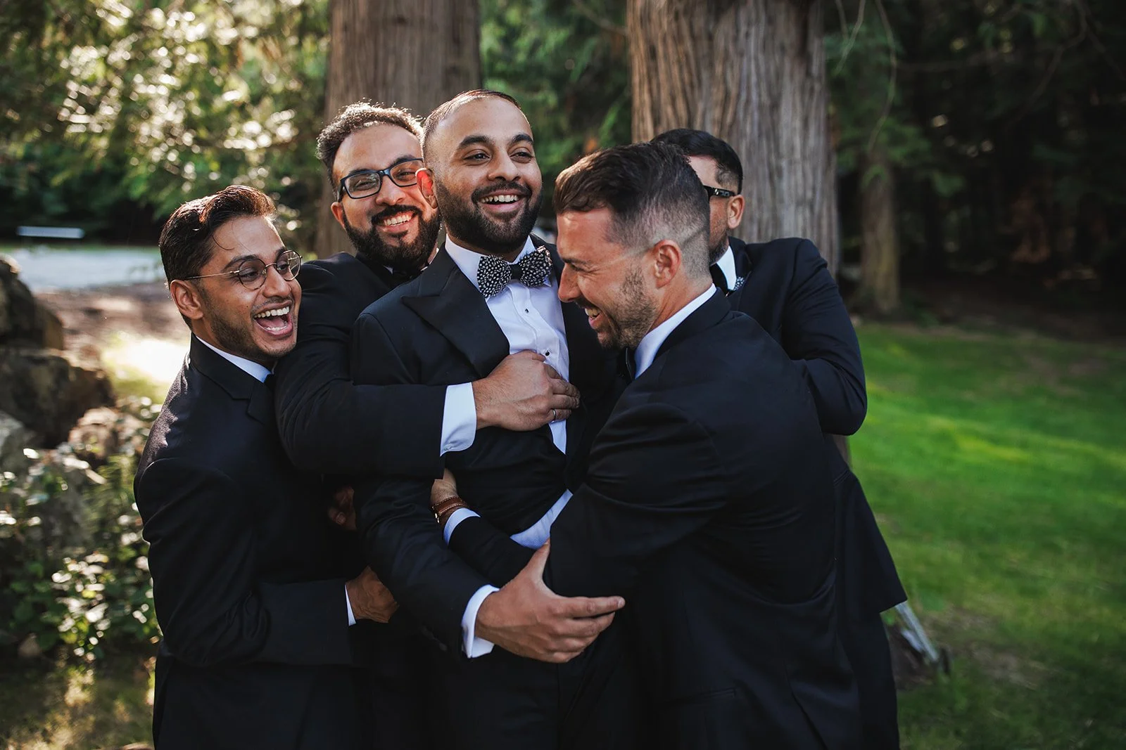 Group of men in tuxedos smiling and hugging outdoors near trees and a pond.