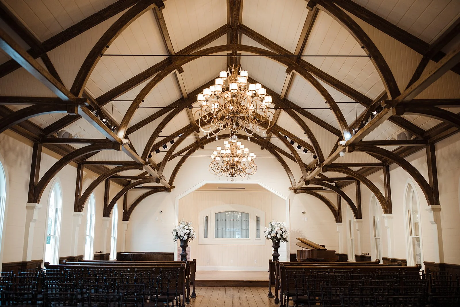 Interior of a decorated wedding or event venue with high arched wooden beams, chandeliers, floral arrangements, and a grand piano.