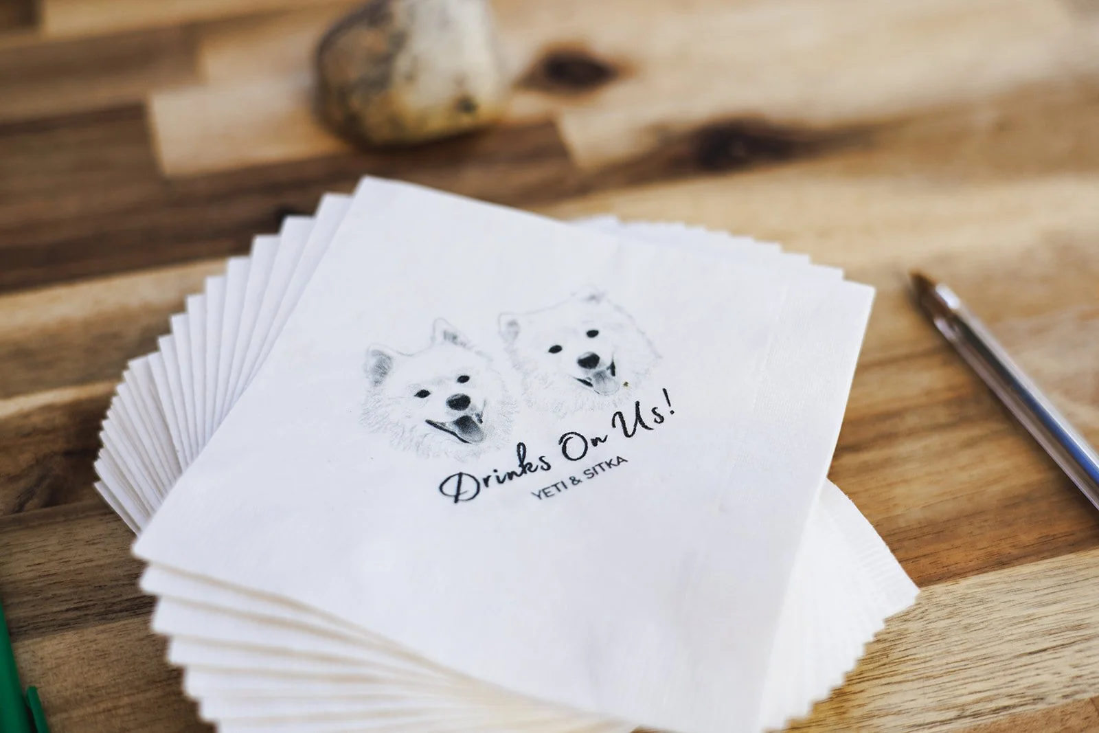 A set of white paper napkins with a sketch of two happy Siberian Huskies and the text 'Drinks Or Us! YETI & SITKA' printed on them, placed on a wooden table.