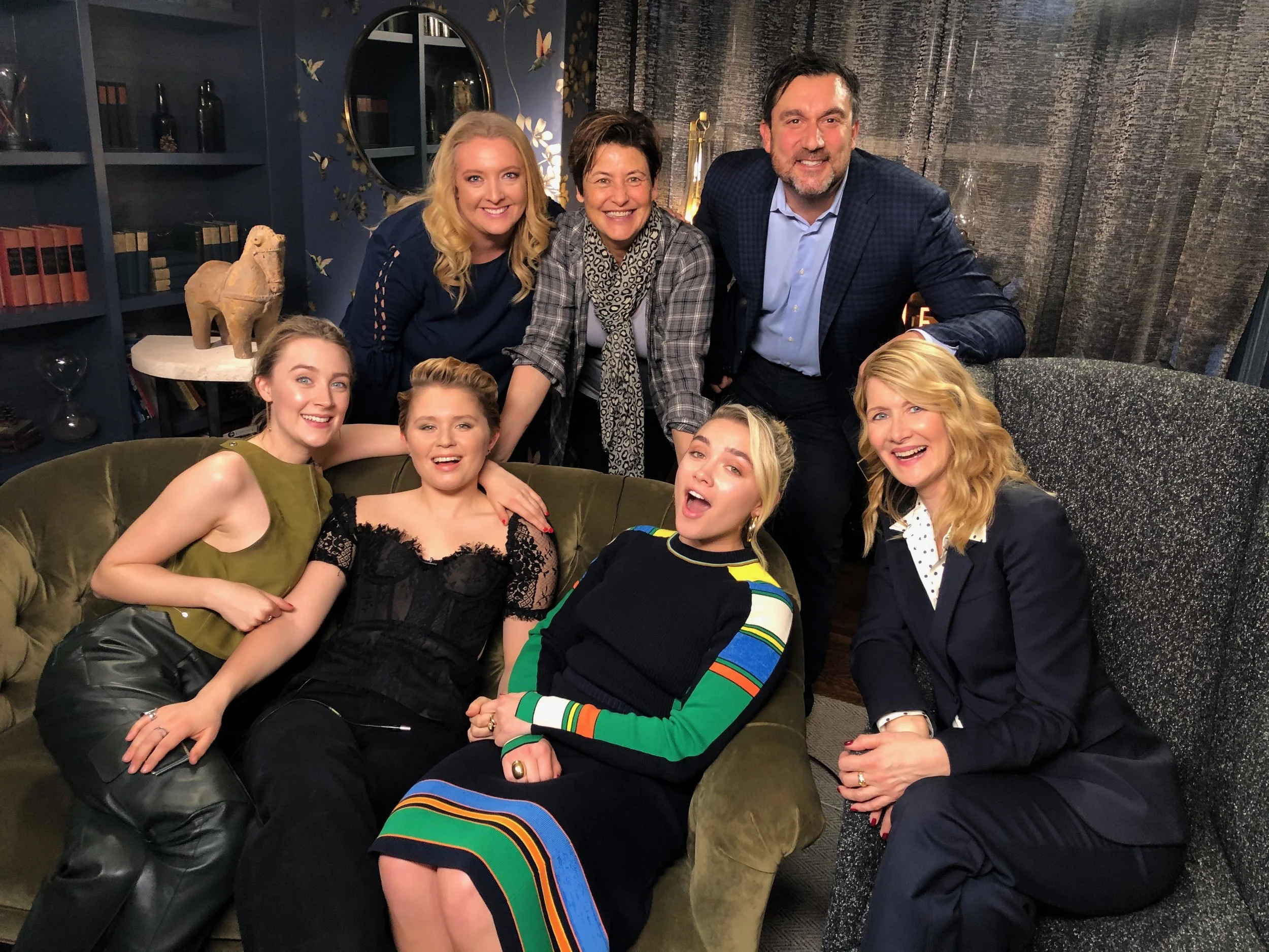 Group of seven people, five women and two men, smiling and posing together in a cozy living room or lounge area. They are seated and standing around a green velvet couch, with decorative shelves and a mirror in the background.