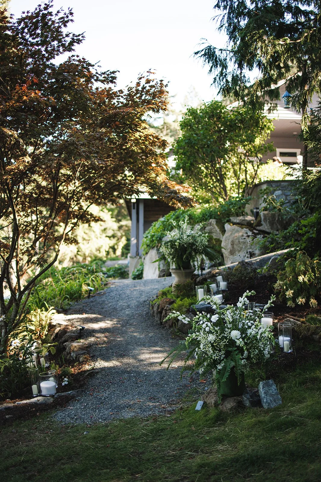 A garden pathway lined with white candles and floral arrangements, leading up to a house with trees and greenery.