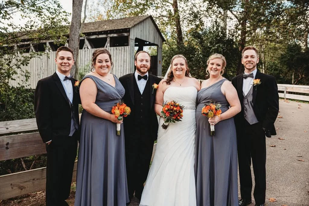 Wedding party with bride in white dress, groom in black tuxedo, and four bridesmaids and groomsmen, outdoors near a rustic barn.