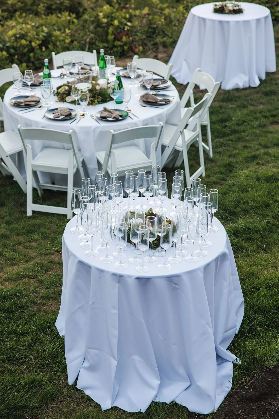 A round outdoor dining table with a white tablecloth set for a meal, surrounded by white chairs. A rectangular table in the foreground is decorated with empty champagne flutes and candles. A smaller round table with a white tablecloth is in the backg