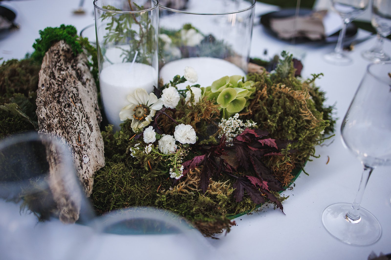 A decorative centerpiece of white candles, flowers, moss, and leaves on a table.
