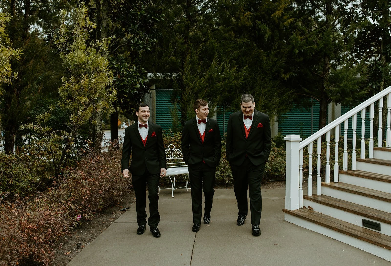 Three men in formal black suits with red accents walking outdoors near a staircase and greenery, smiling and talking.