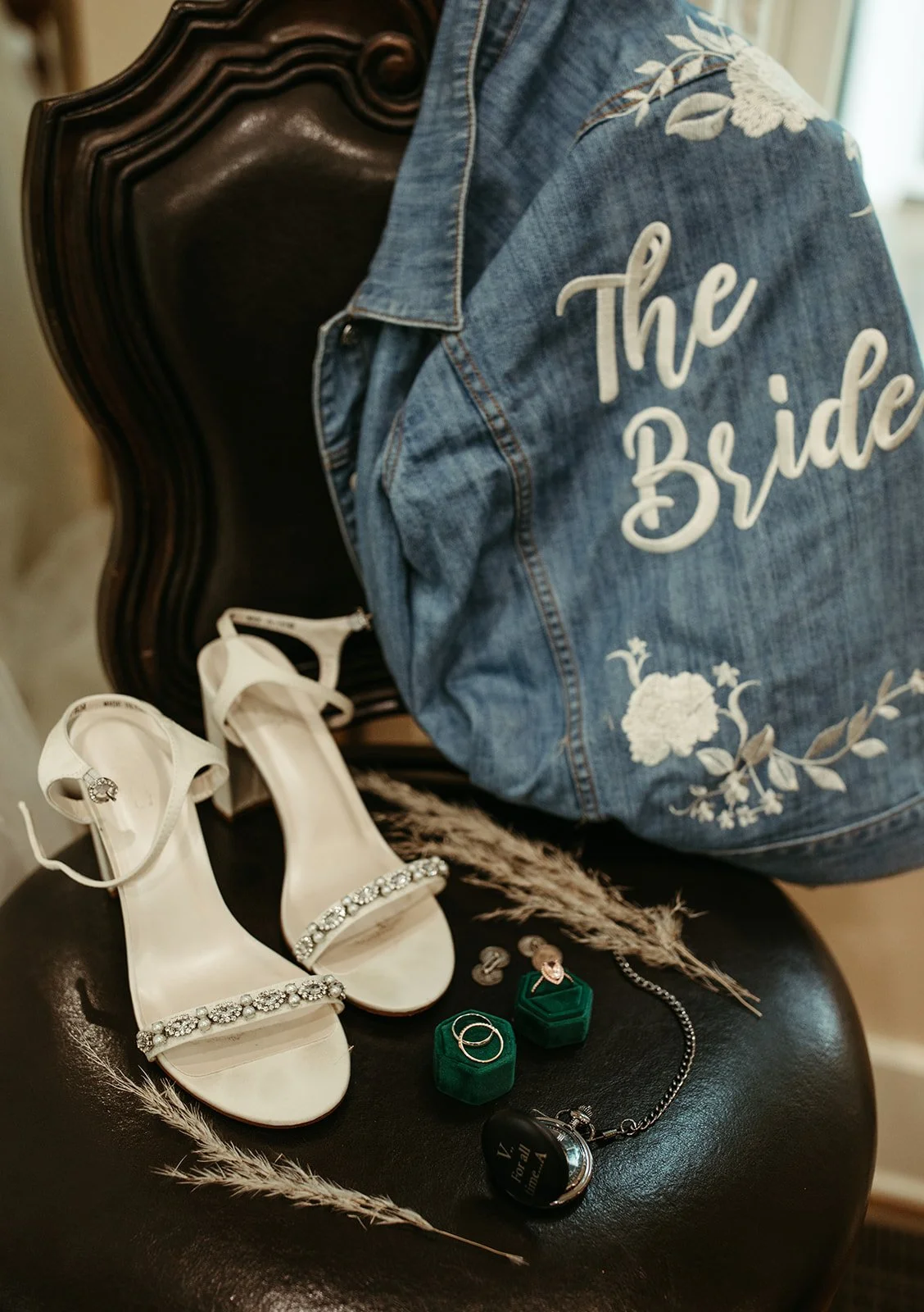 Detail of a wedding or bridal preparation scene, showing a denim jacket with 'The Bride' written on the back, a pair of white embellished high-heeled sandals, green velvet ring boxes, jewelry including rings and earrings, a watch, and decorative dried plants on a black surface.