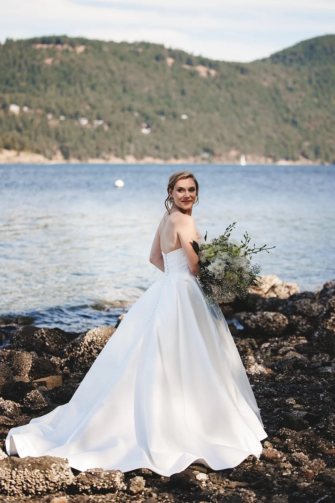 Bride in a white wedding gown holding a bouquet standing on rocky shoreline by a lake with mountains in the background.
