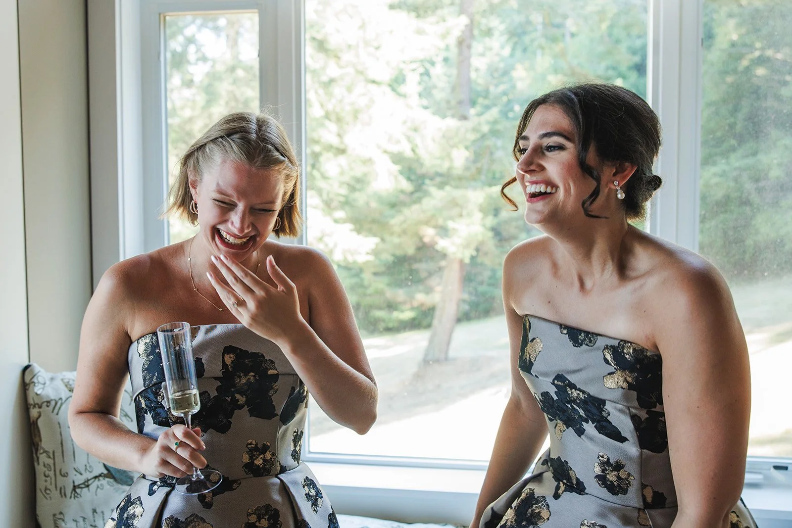 Two women in strapless floral dresses laughing and sharing a moment together, one holding a glass of champagne, in front of a large window with a view of trees.