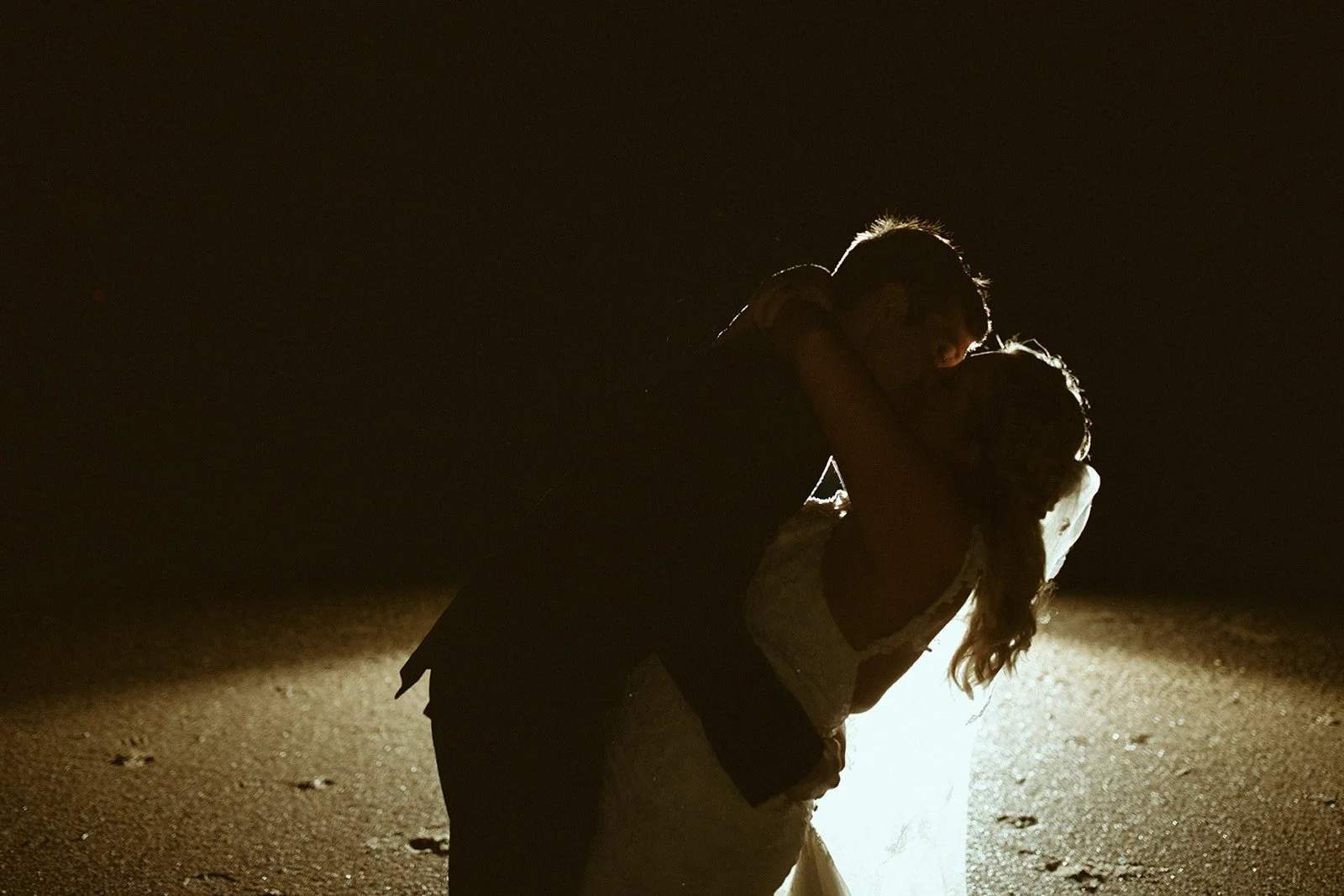 Silhouettes of a couple kissing on a beach at night, backlit by a bright light, with the sandy ground visible.