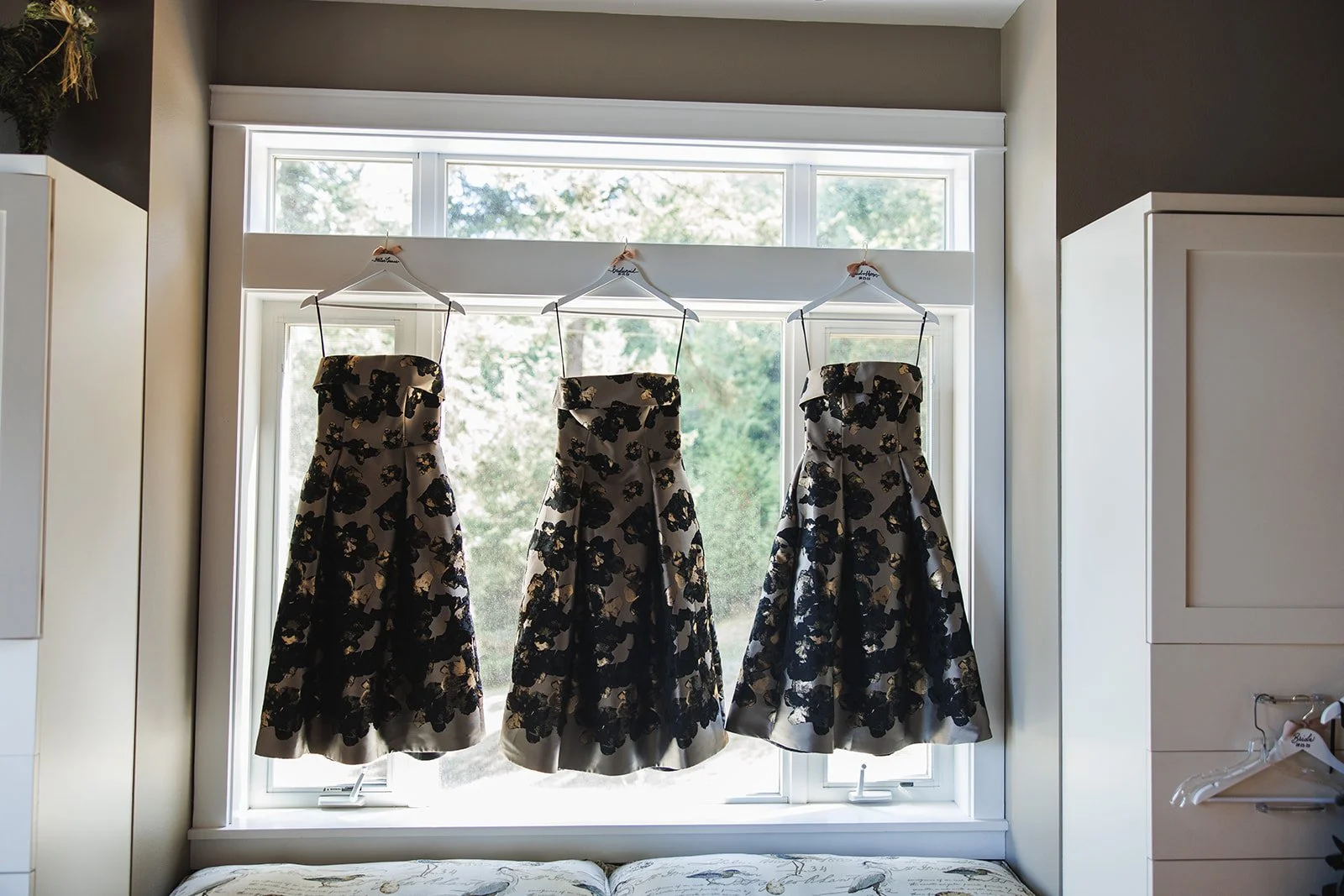 Three matching black and beige floral dresses hanging on hangers in front of a large window.