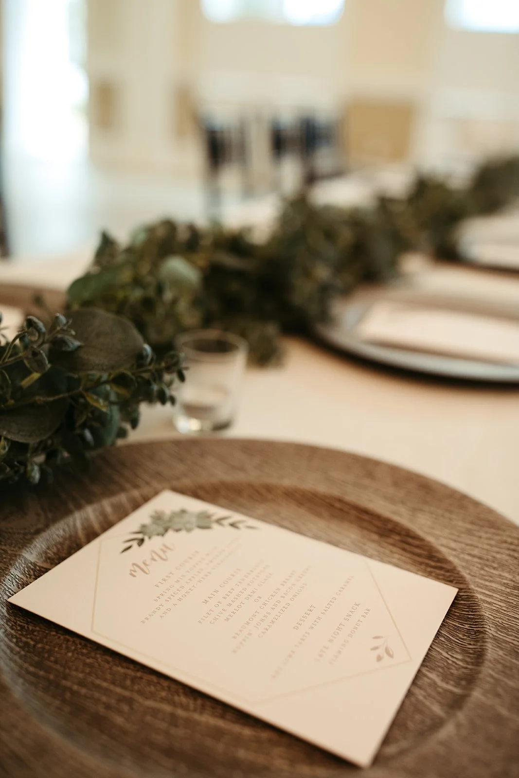 Close-up of a wedding menu on a dark wooden plate, decorated with greenery and a blurred dining table in the background.