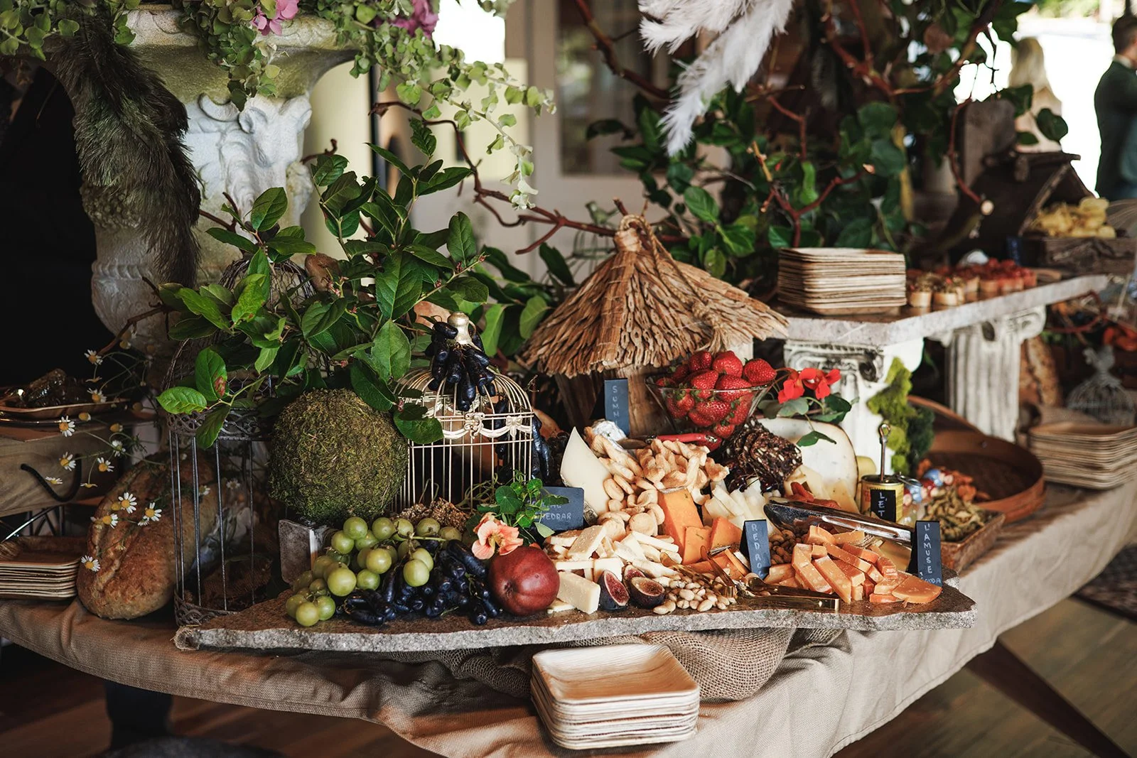 A cheese, fruit, and snack display on a table at a buffet or party, with cheeses, grapes, strawberries, figs, chips, and decorative greenery and flowers.
