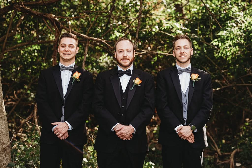 Three men dressed in black tuxedos with bow ties, standing outdoors in front of trees, each wearing a boutonniere with a peach-colored flower.