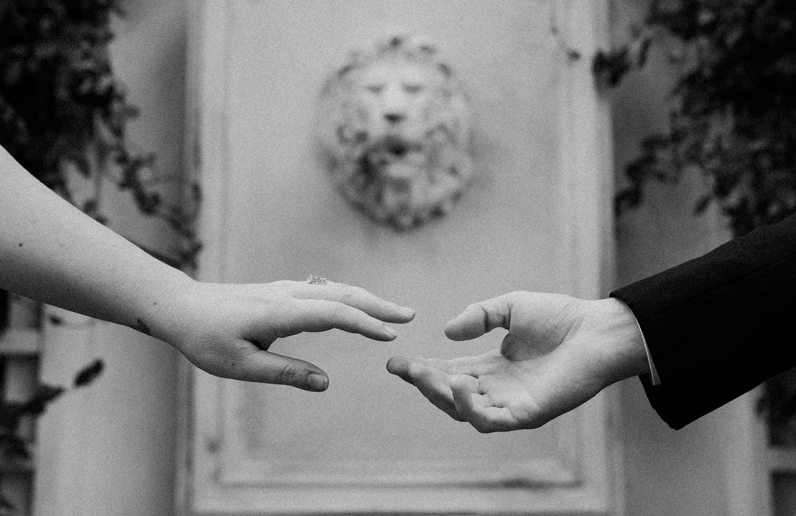Black and white photo of two people reaching out to each other with their hands, with a lion face sculpture and a wall with plants in the background.