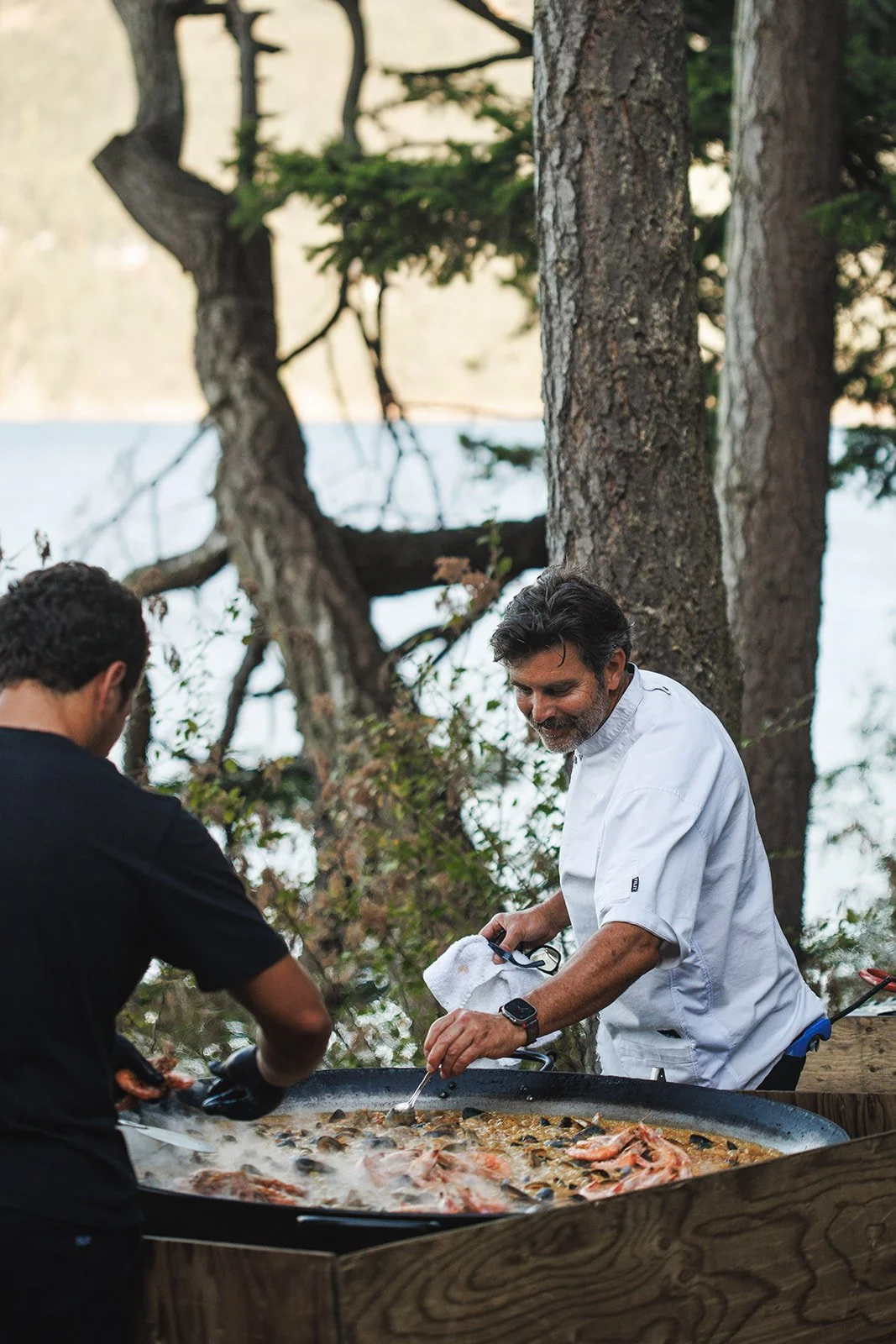 Two men cooking paella outdoors in a large pan surrounded by trees near a lake.