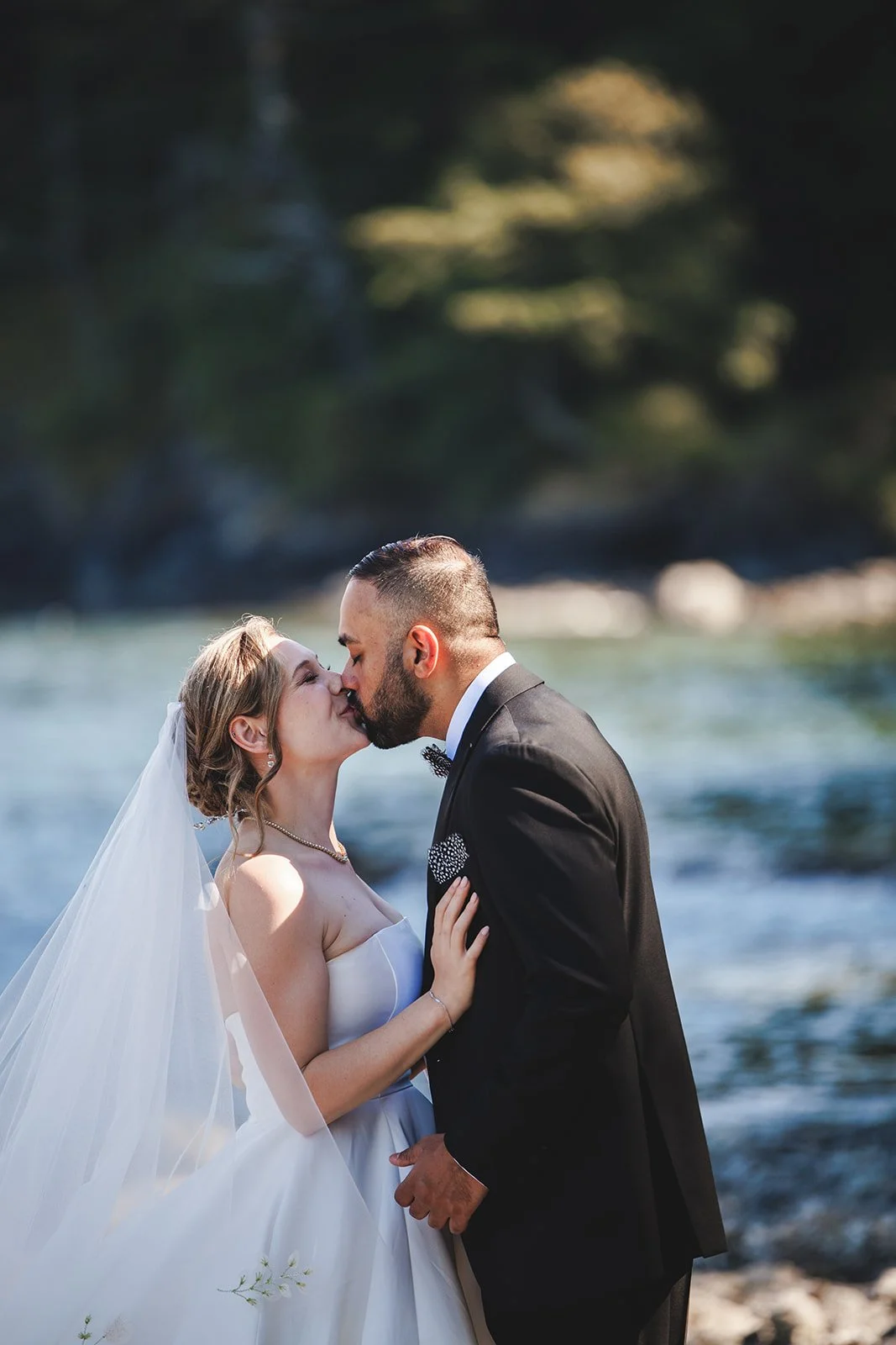 A bride and groom are kissing outdoors near a river, with trees in the background.