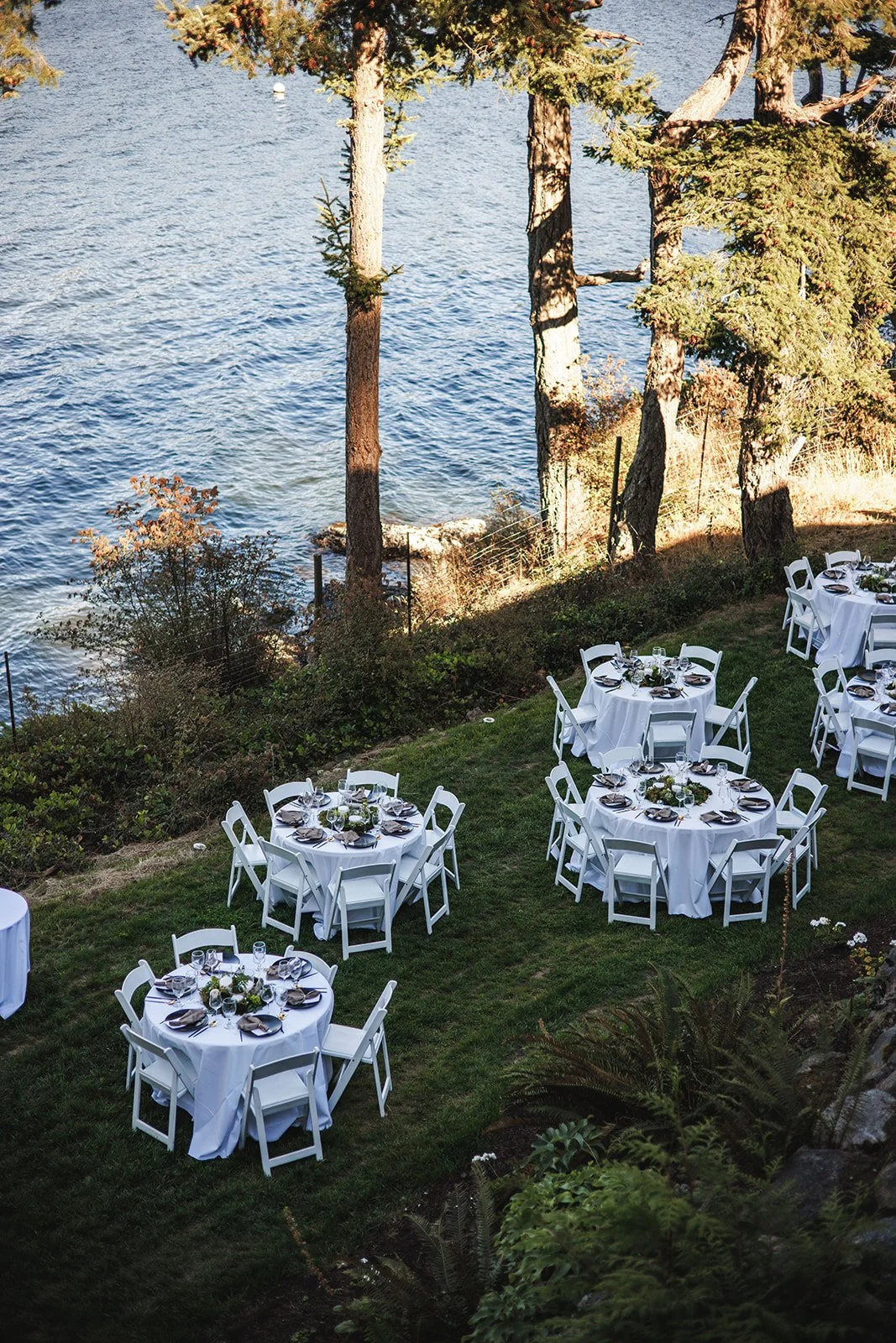 Outdoor dining tables with white tablecloths and chairs set up on grass near a lake with trees along the shore.