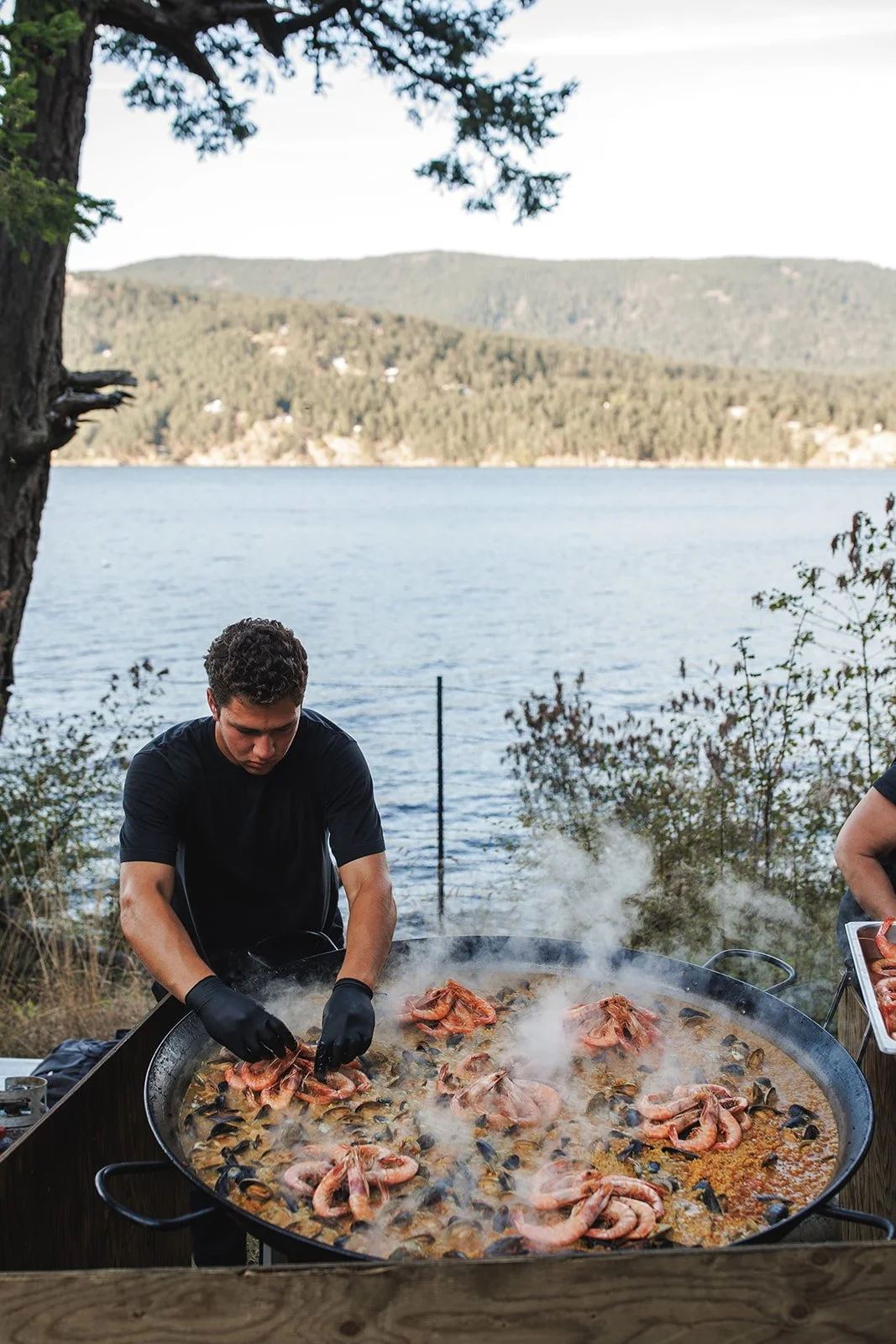 A man wearing black gloves is cooking a large seafood paella with shrimp, mussels, and rice outdoors by a lake, with mountains and trees in the background.