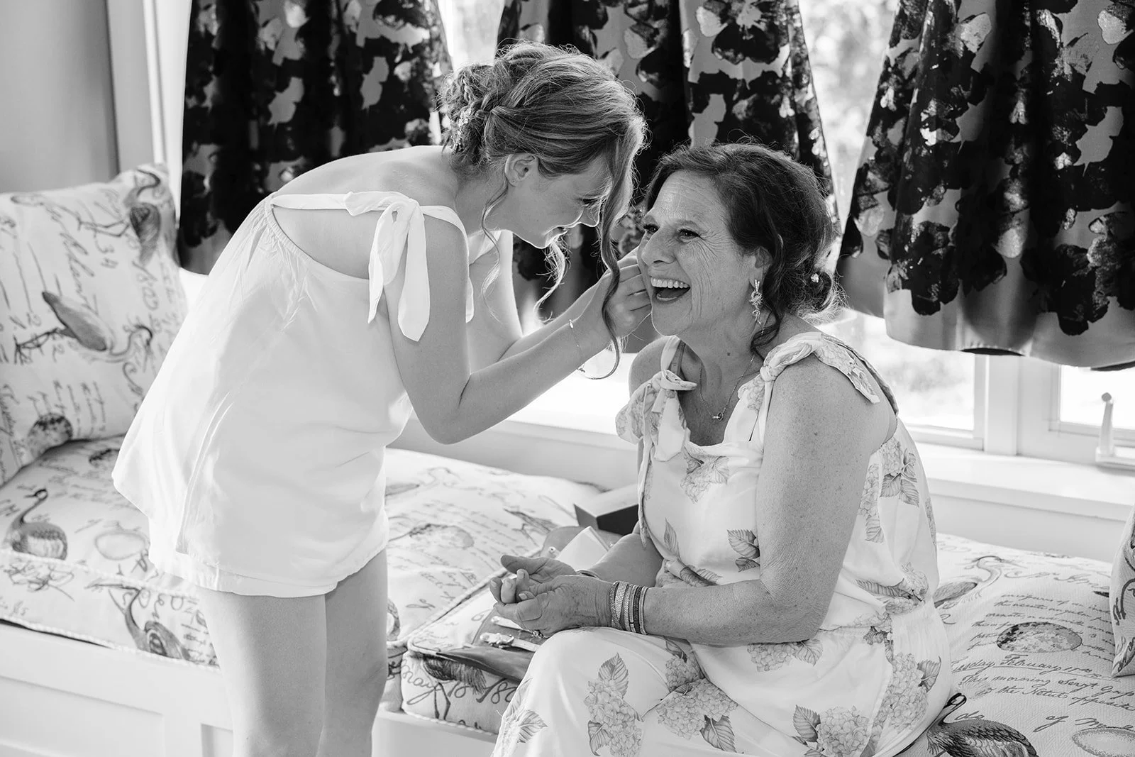 A young woman is applying makeup to an older woman who is sitting on a bed, smiling and enjoying the moment.