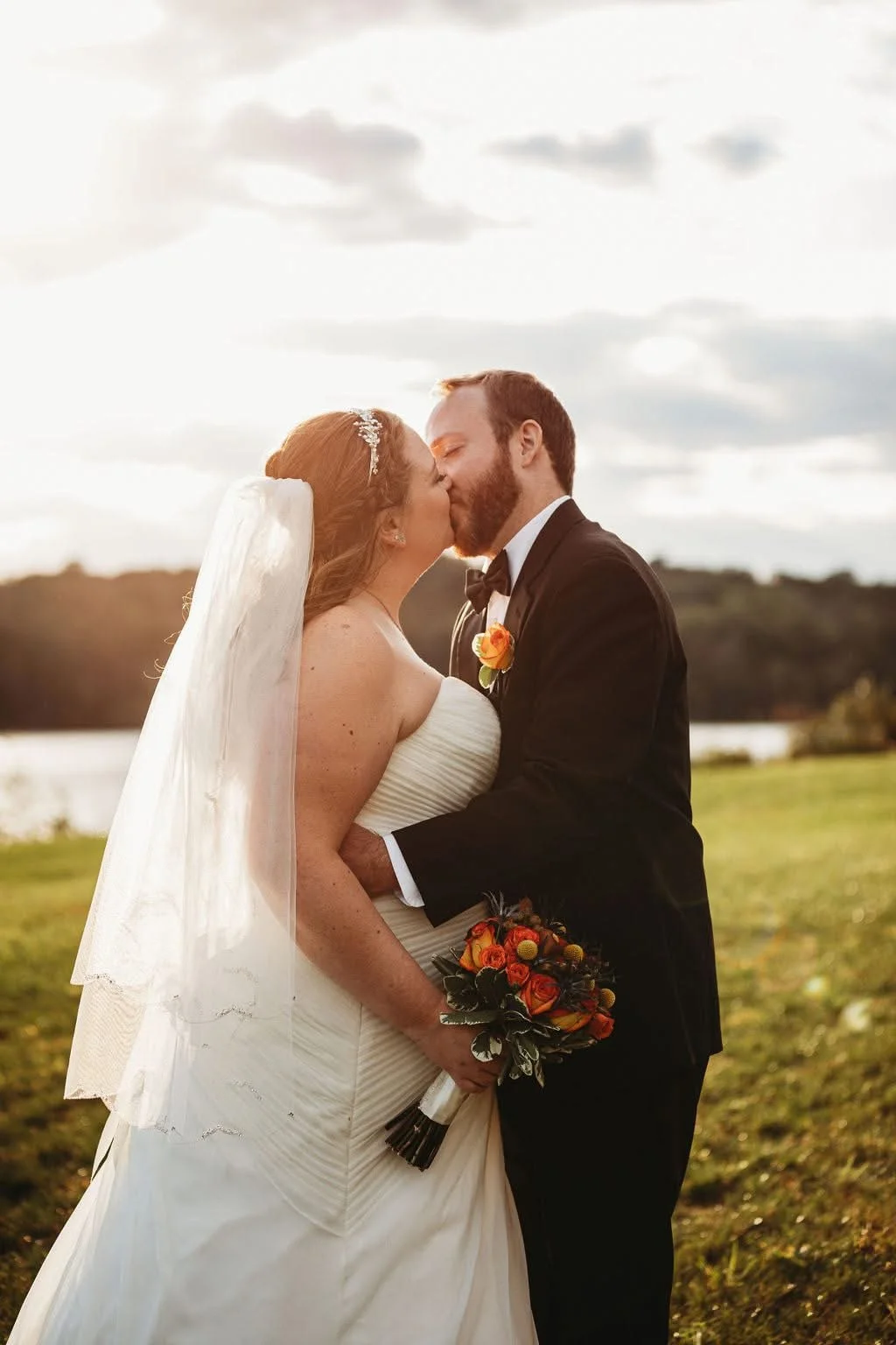 A newlywed couple sharing a kiss outdoors during sunset, the bride in a white wedding dress and veil holding a bouquet of orange and yellow flowers, the groom in a black tuxedo with a yellow boutonniere, with a scenic landscape and cloudy sky in the 