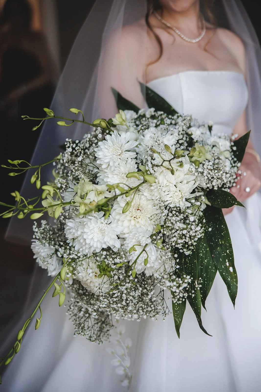 Bride in a white wedding dress holding a large bouquet of white flowers, including dahlias, baby's breath, and lilies, with green leaves, wearing a pearl necklace and veil.