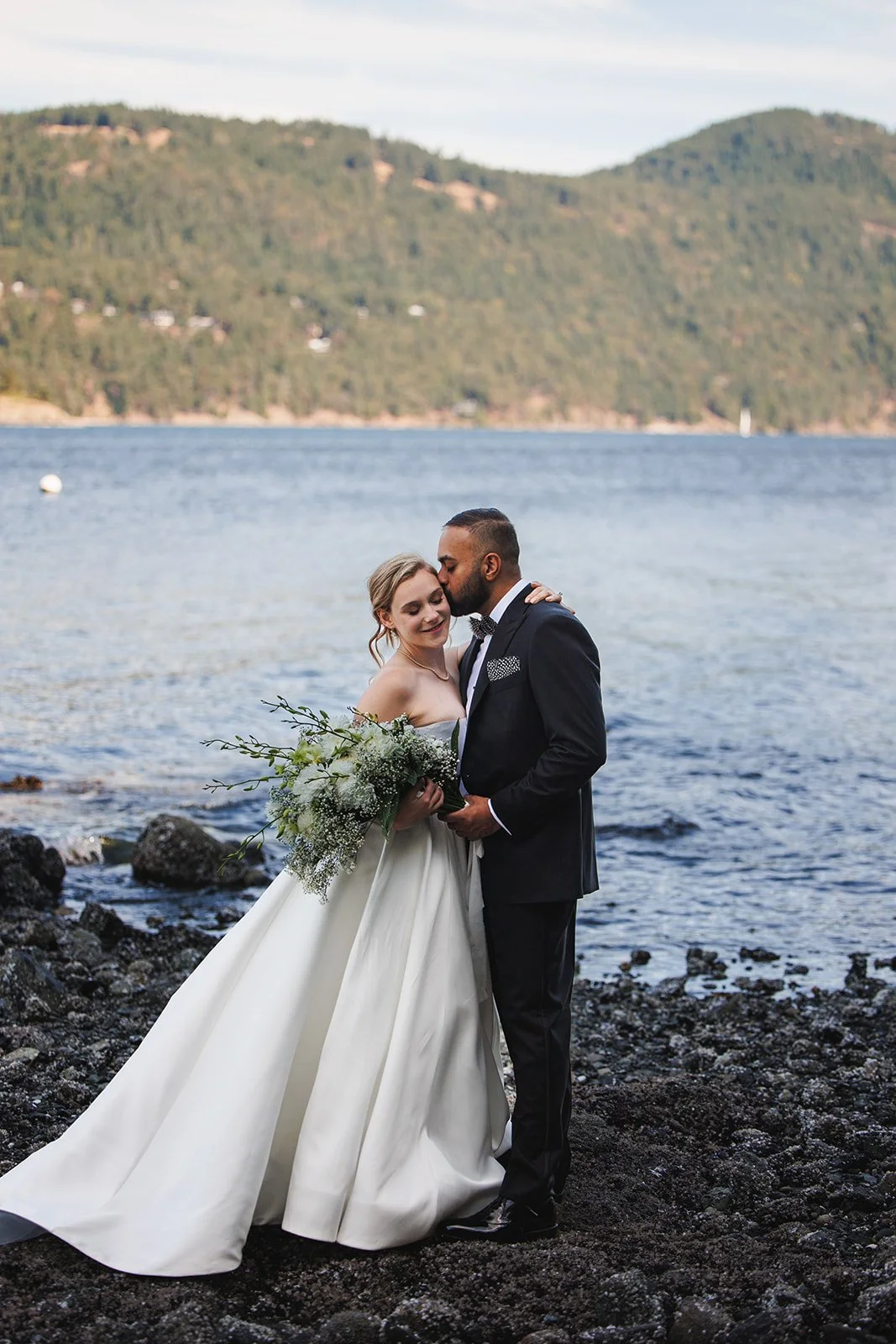 A bride and groom stand on a rocky shore by a lake, embracing. The bride is in a strapless white wedding gown holding a bouquet of white and green flowers. The groom is in a dark suit with a bow tie, kissing her forehead. Hills and water are visible 