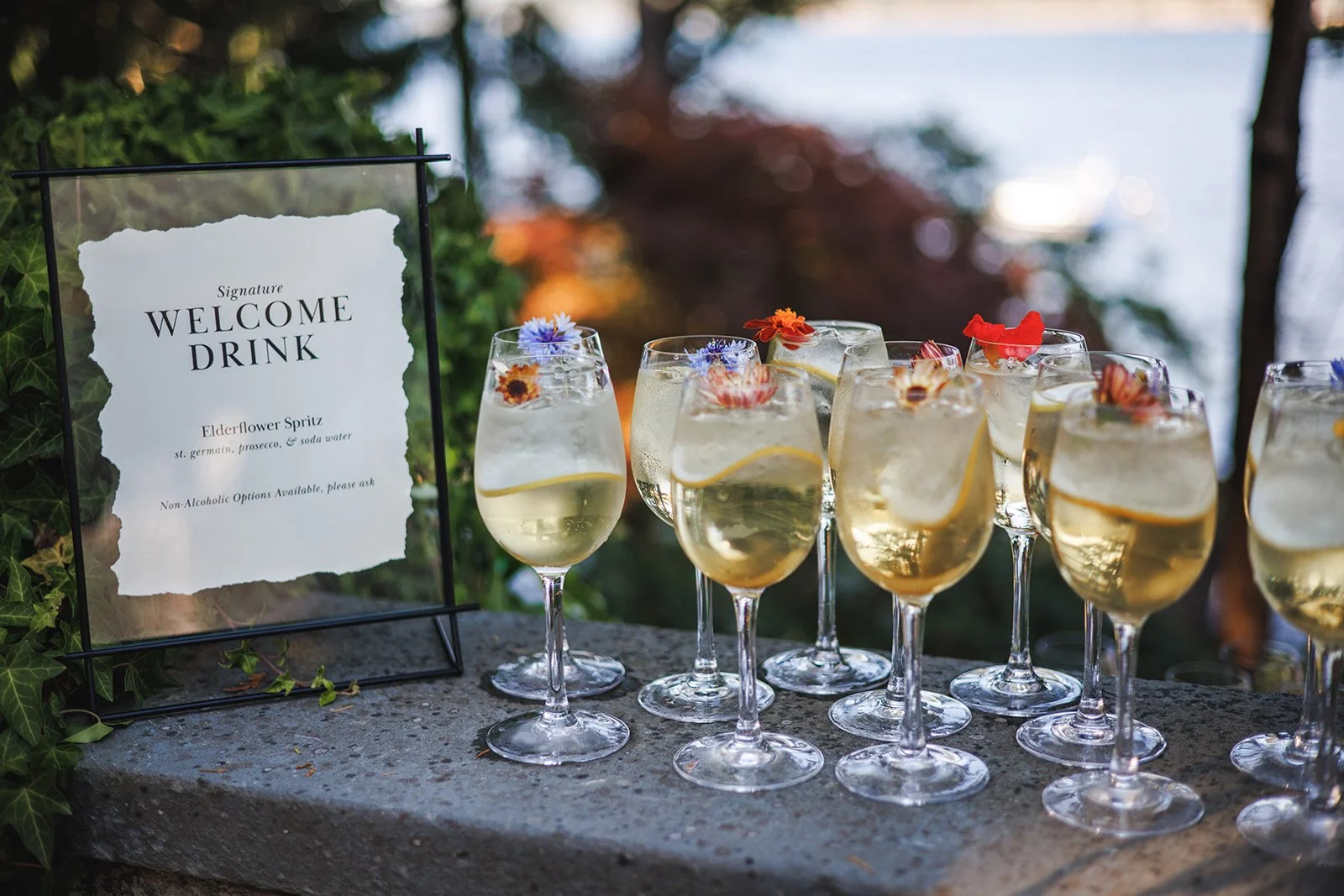 Glass bowls of elderflower spritz drinks garnished with edible flowers on a stone surface at an outdoor event.