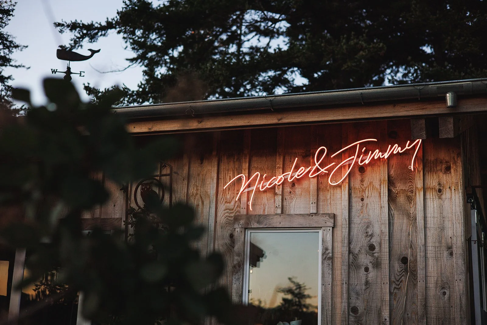 A wooden building exterior with a neon sign displaying the names 'Nicole & Jimmy' in cursive. The sign glows pinkish-orange, and there is a small window below it reflecting the outdoors. The background shows trees and an overcast sky.