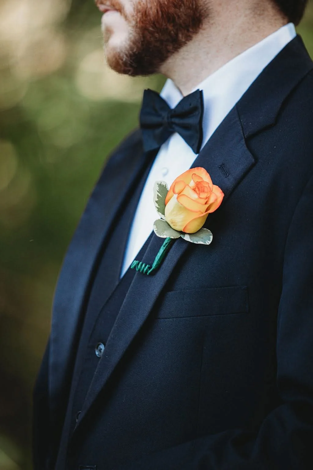 Close-up of a man in a tuxedo with a black bowtie, wearing a flower boutonniere featuring a peach-colored rose and greenery on his lapel.