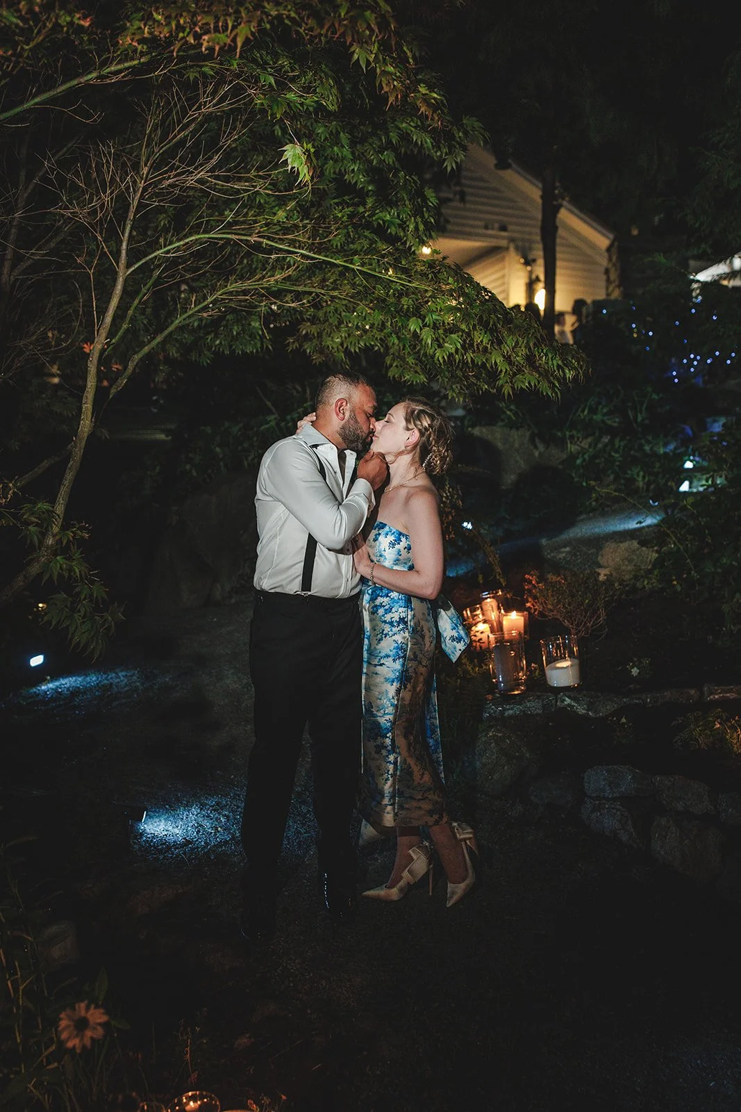 A romantic couple sharing a kiss outdoors at night, surrounded by trees and lanterns, with a house in the background.