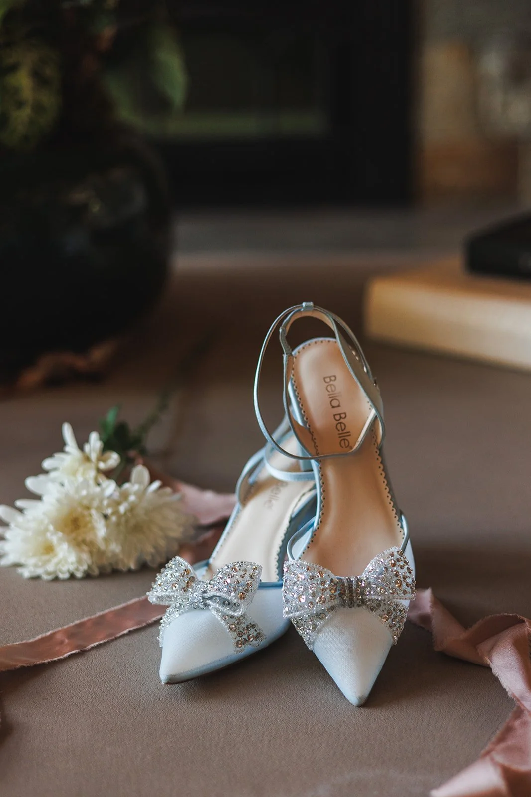 Elegant white bridal shoes with peep-toe design, adorned with sparkling rhinestone bows, placed on a beige surface with a pink satin ribbon and a white flower nearby.