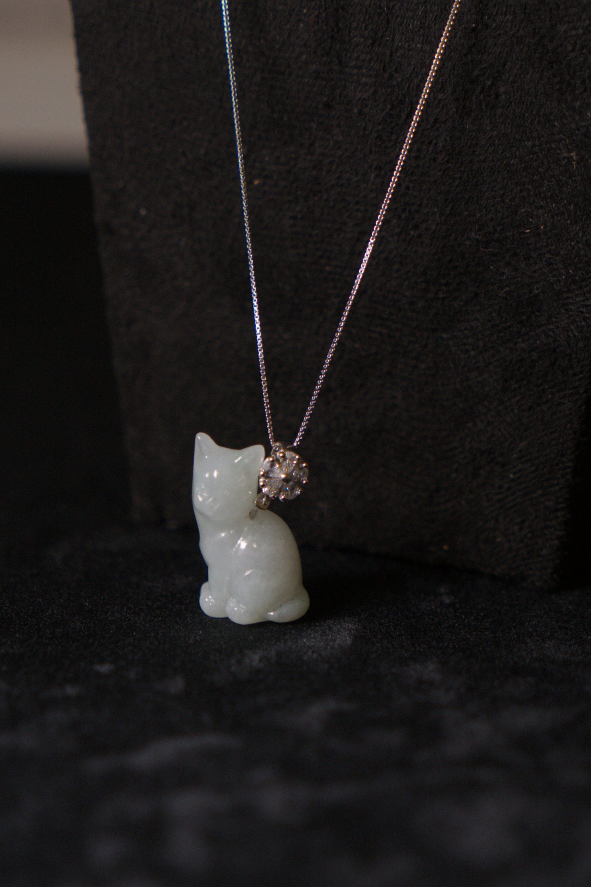 A white jade cat-shaped pendant necklace with a silver and diamond floral charm on a black fabric background.