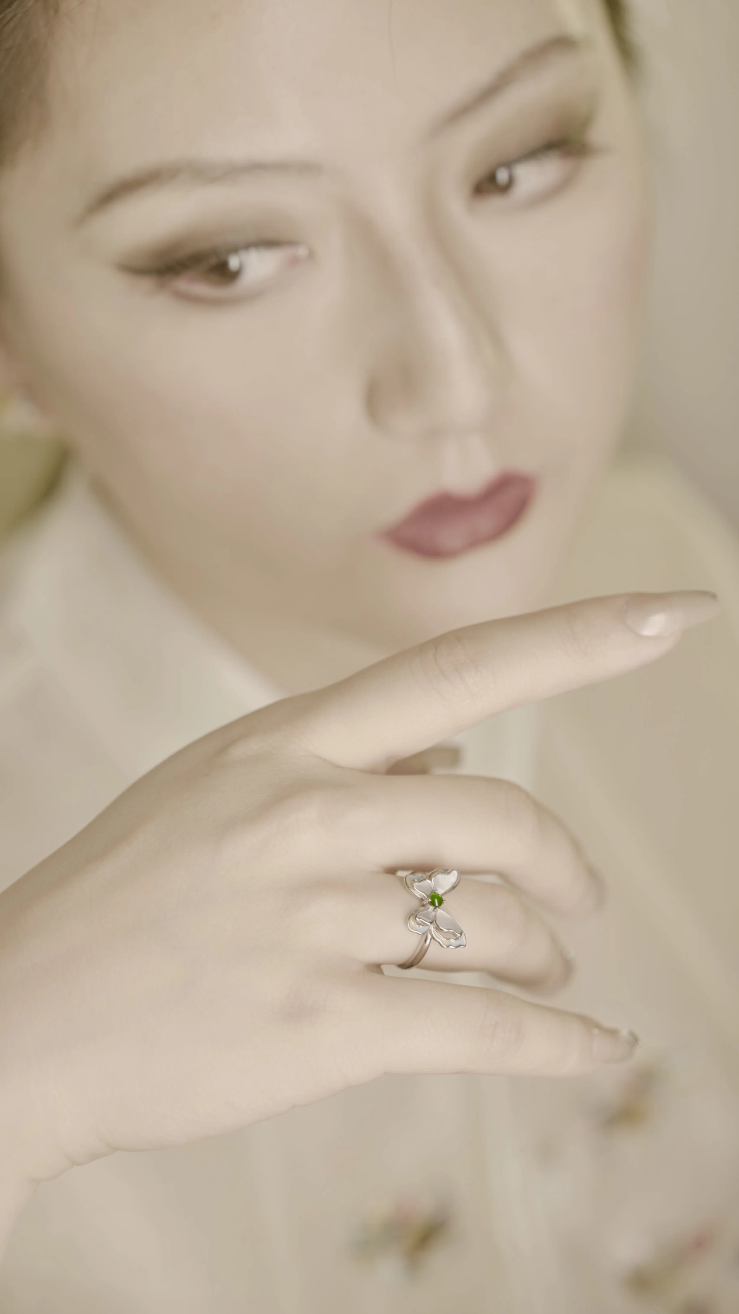 Close-up of a woman with makeup looking down, showing her hand with a butterfly-shaped silver ring with a green jadeite stone.