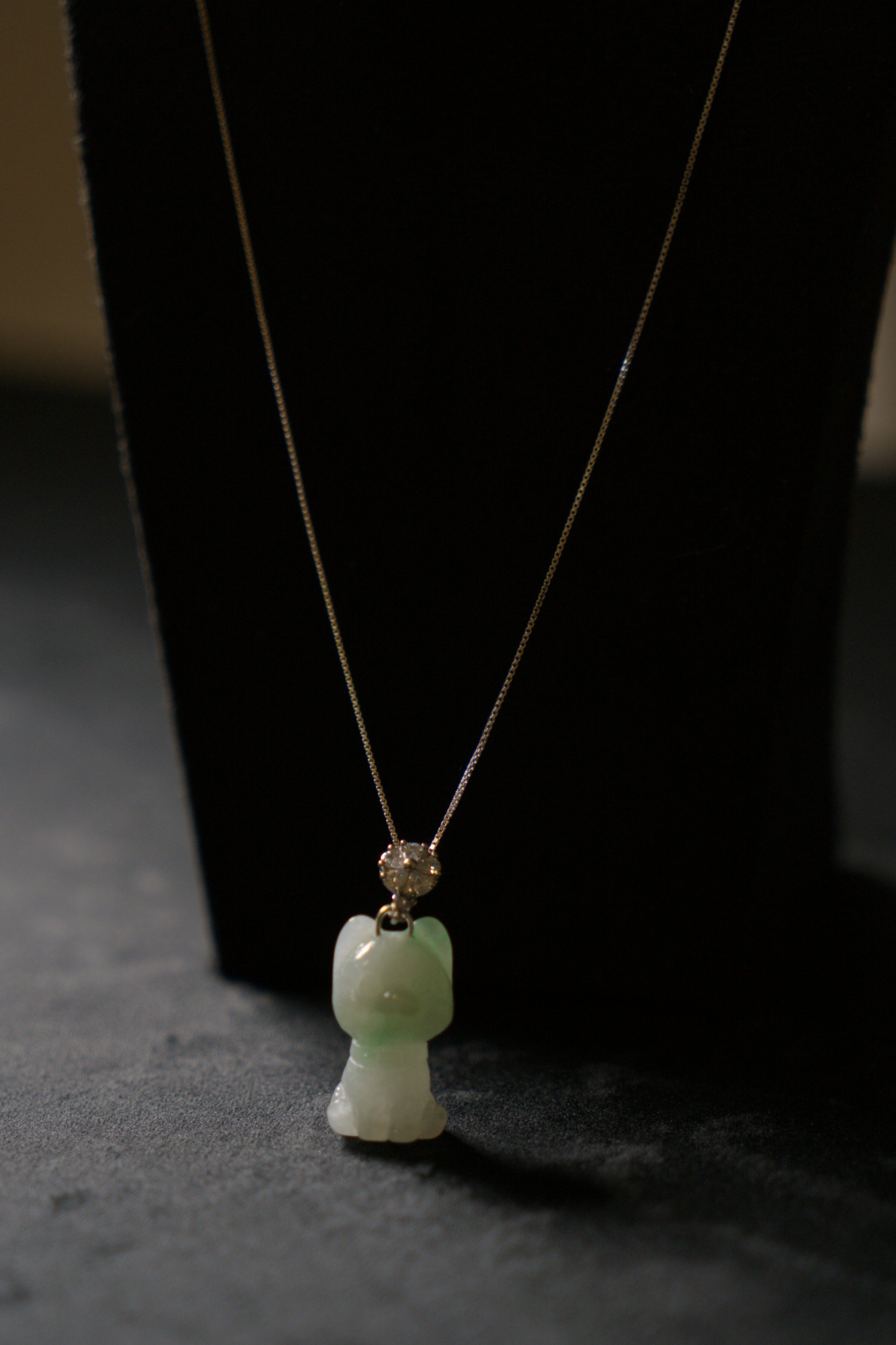 Jade necklace with a carved jade bear pendant and a small gemstone in a silver setting, displayed on a black stand.
