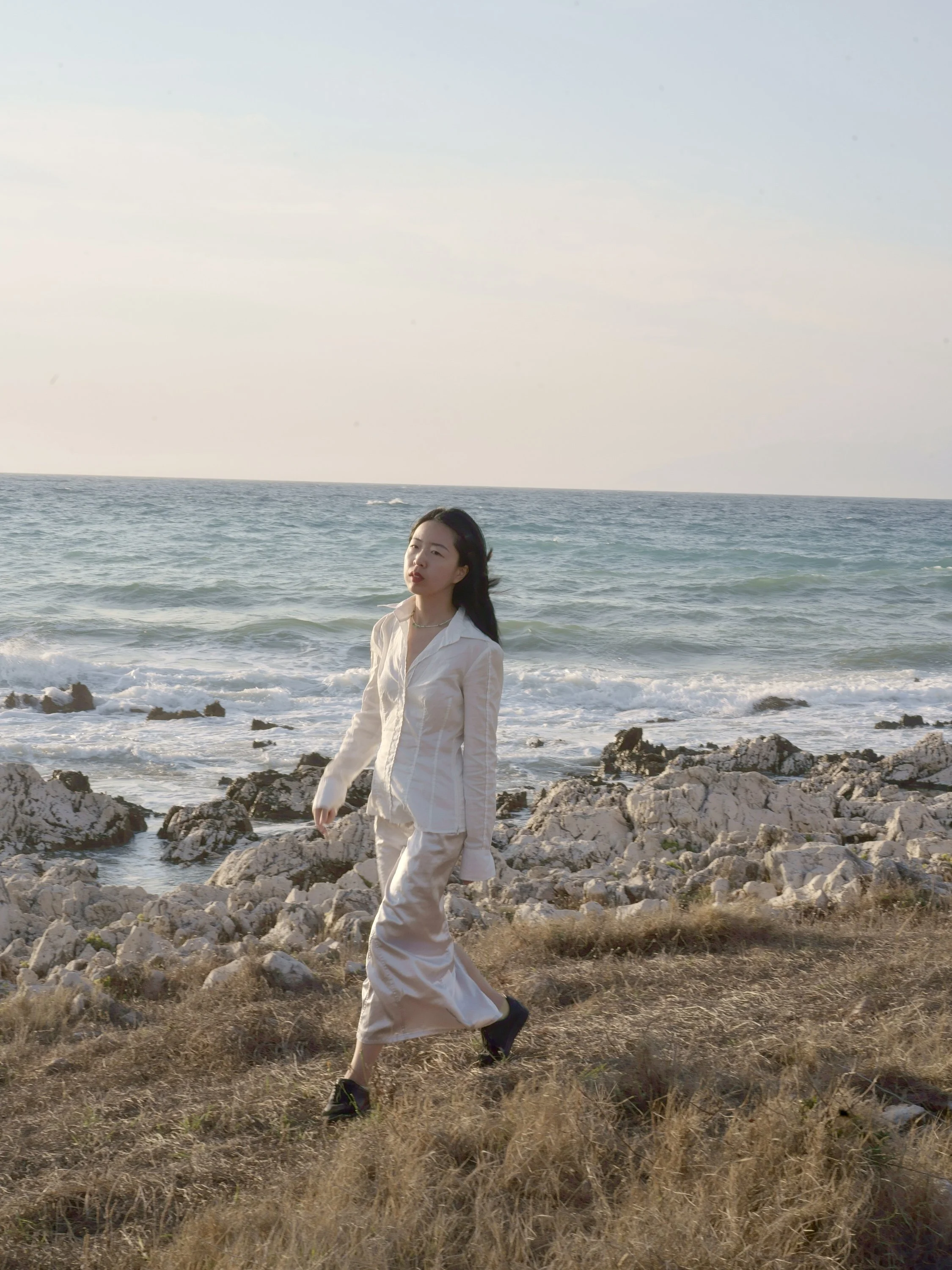 A woman in a white satin outfit walking on a rocky, grassy shoreline with the ocean and a clear sky in the background.