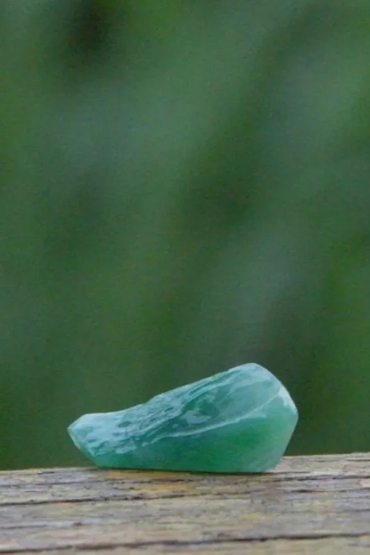 A small, polished green stone resting on a weathered wooden surface with a blurred green background.