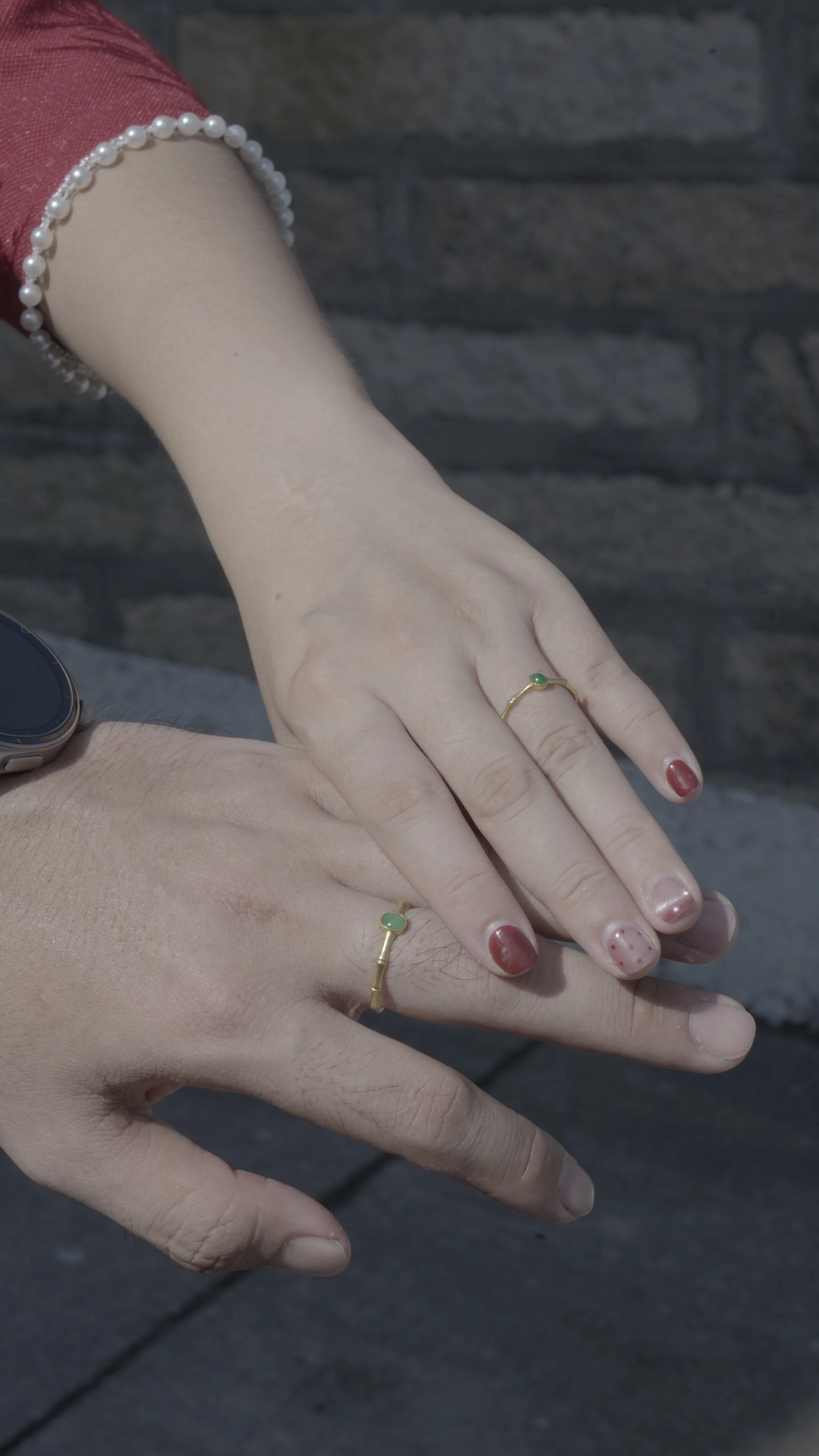 Two hands, one with a ring with a green stone and a bracelet made of pearls, the other with a similar ring, resting on a dark stone surface.