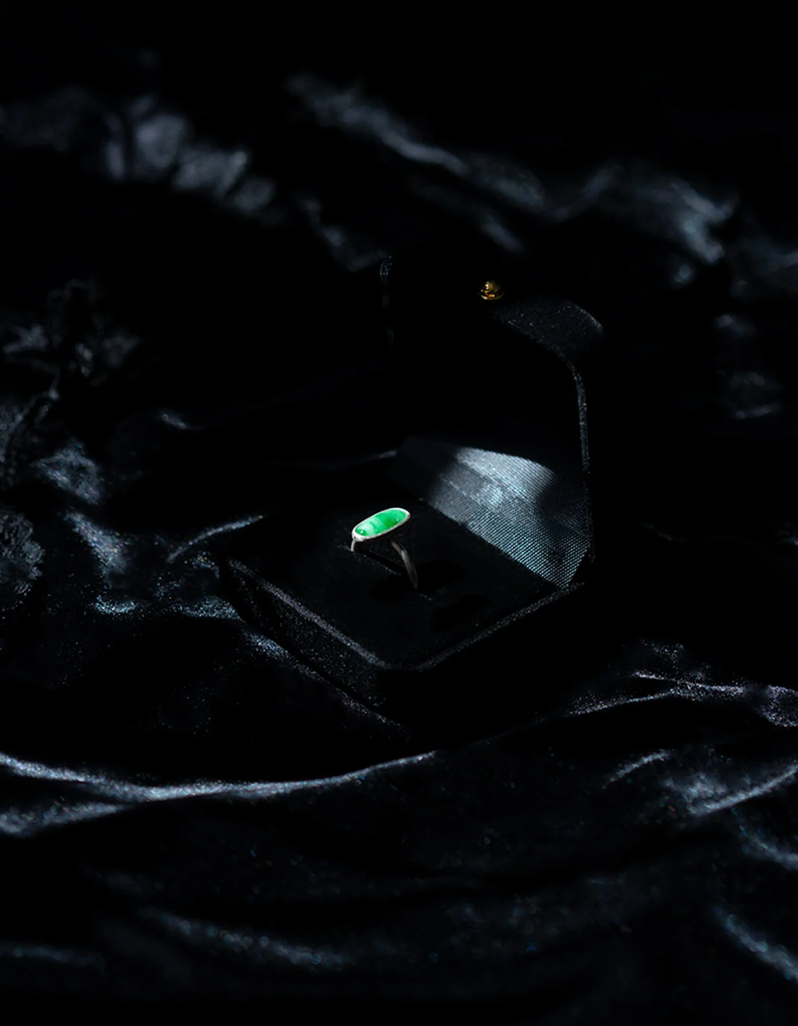 A silver ring with a green stone displayed inside a black velvet box, resting on black satin fabric.