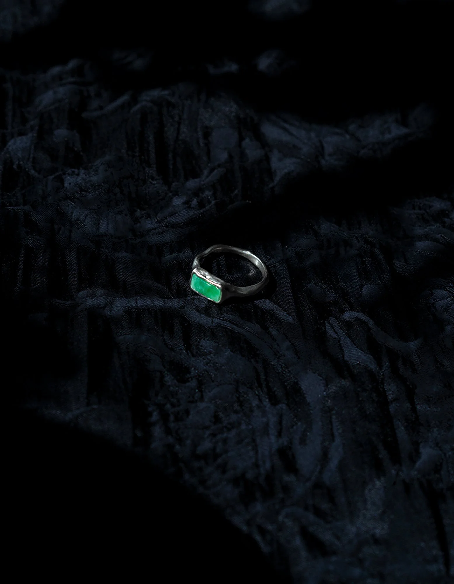 Silver ring with a rectangular green gemstone resting on black textured fabric.