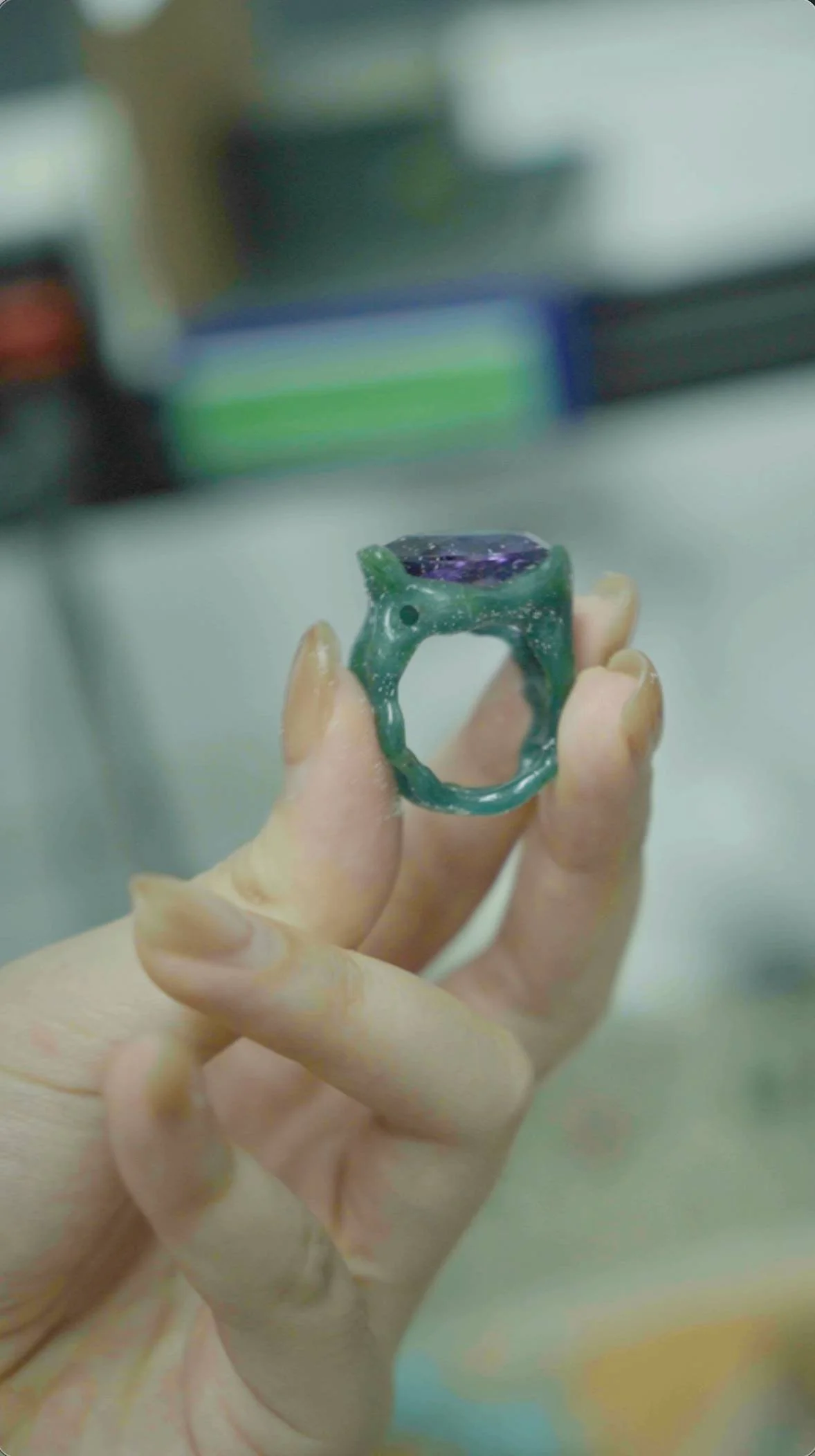 A close-up of a hand holding an intricate, handcrafted ring with a large purple gemstone, in a workshop or studio setting.