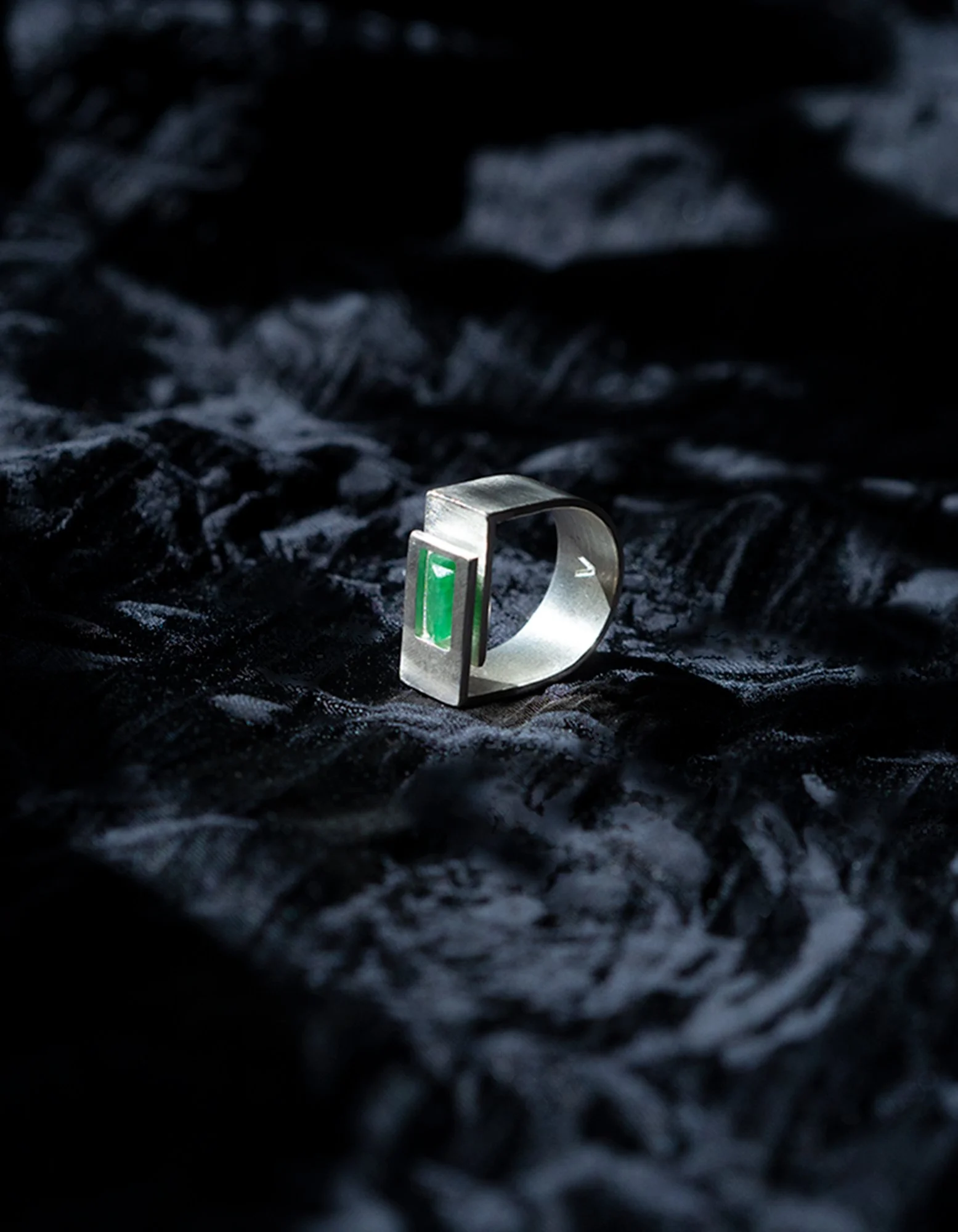 A silver ring with a rectangular green gemstone on a crinkled black fabric background.