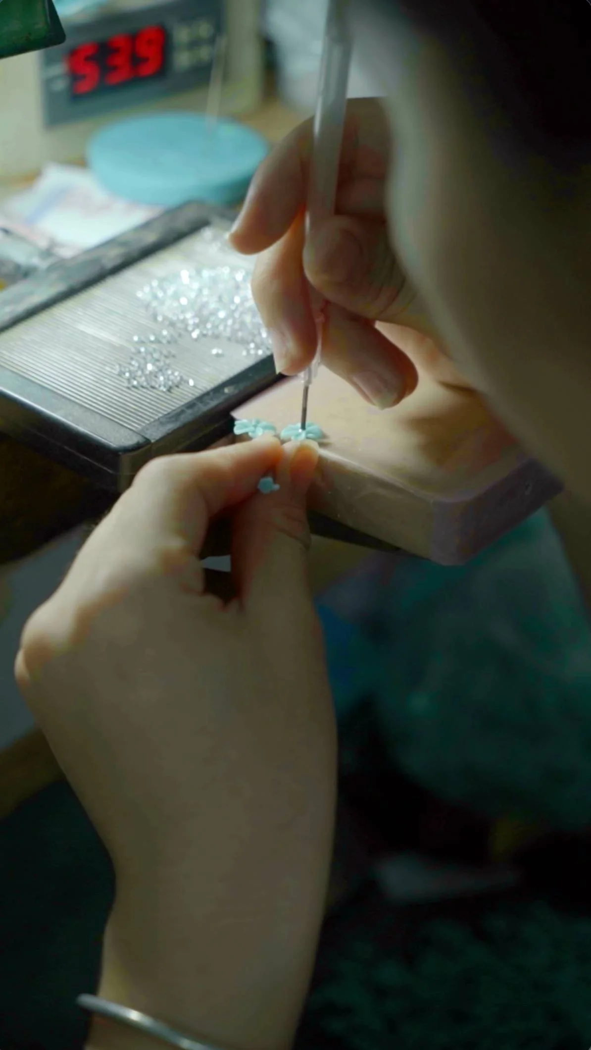 Person using a jewelry beading tool to place small blue flower-shaped beads onto a string, with a tray of jewelry-making supplies nearby.