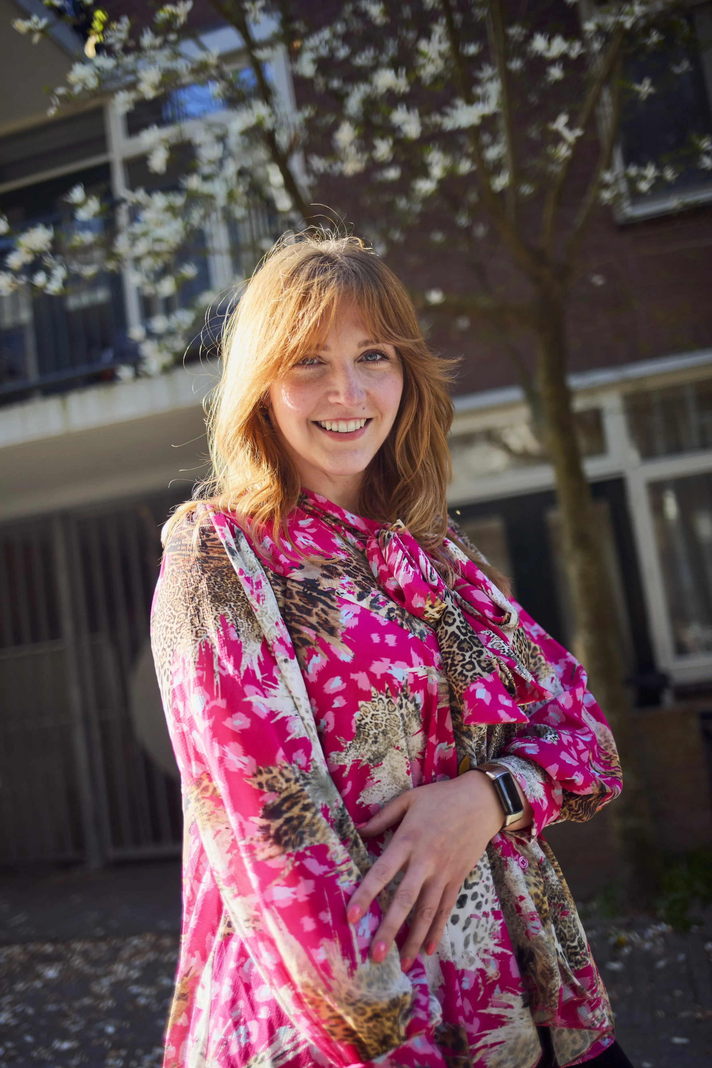 A woman with red hair smiling and looking at the camera outdoors on a sunny day, standing in front of a flowering tree and a building.