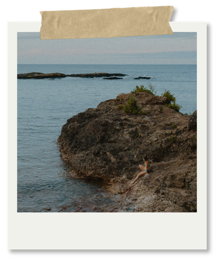 A person sitting on large rocks by the ocean with small islands in the background, under a partly cloudy sky.