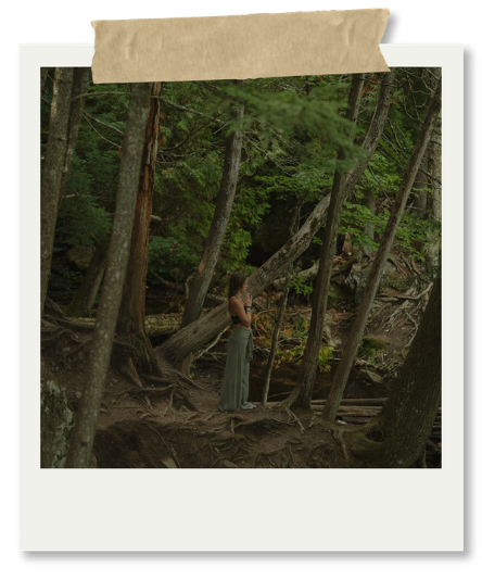 A woman standing on a forest floor among trees and roots, with her hands covering her face, in a wooded area.