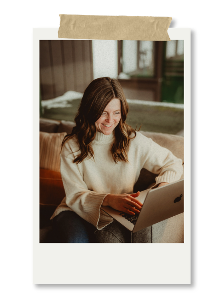 Woman with brown hair smiling while using a laptop, sitting on a couch in a cozy indoor setting.