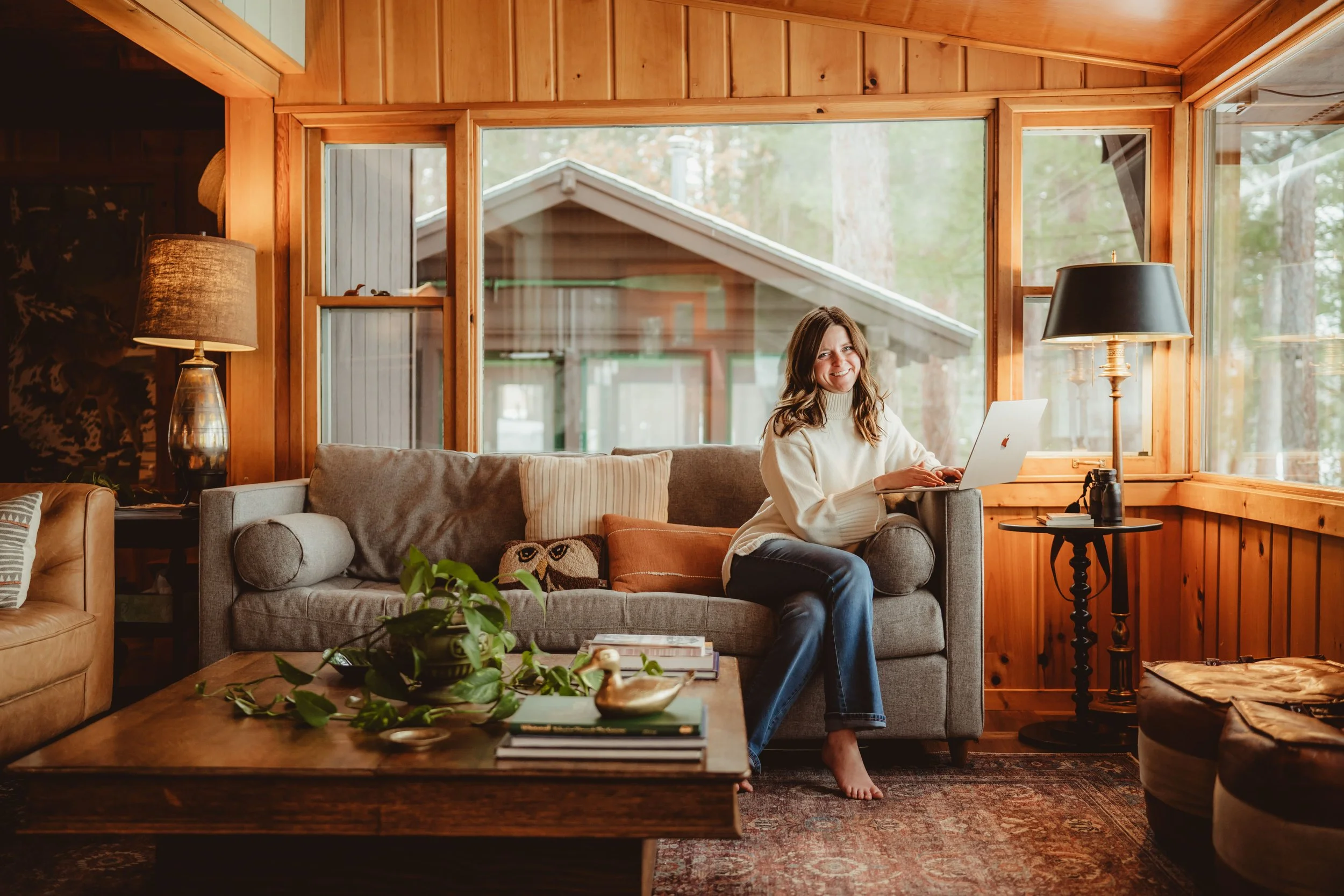 A young woman with brown hair sitting on a grey sofa in a wooden cabin living room, smiling at the laptop on her lap, with large windows behind her showing trees outside.