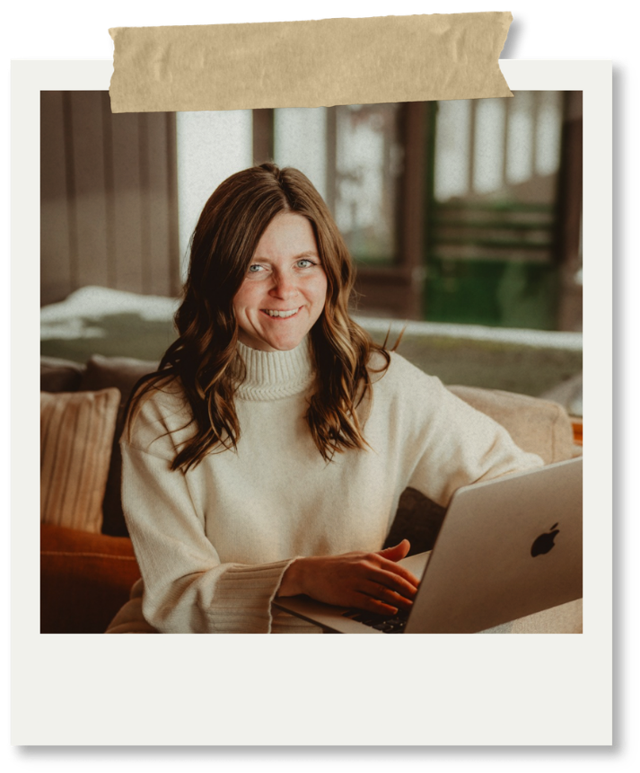 A woman with long brown hair and a cream-colored sweater sitting in a cozy room with a laptop, smiling and looking at the camera.