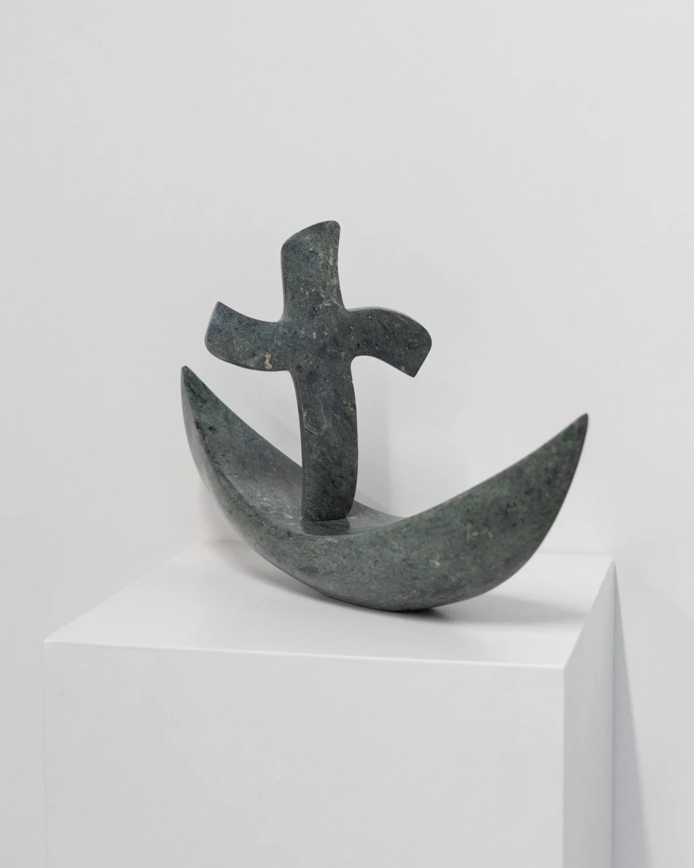 A modern abstract sculpture of a cross and a crescent shape, made of stone, displayed on a white pedestal against a plain white background.