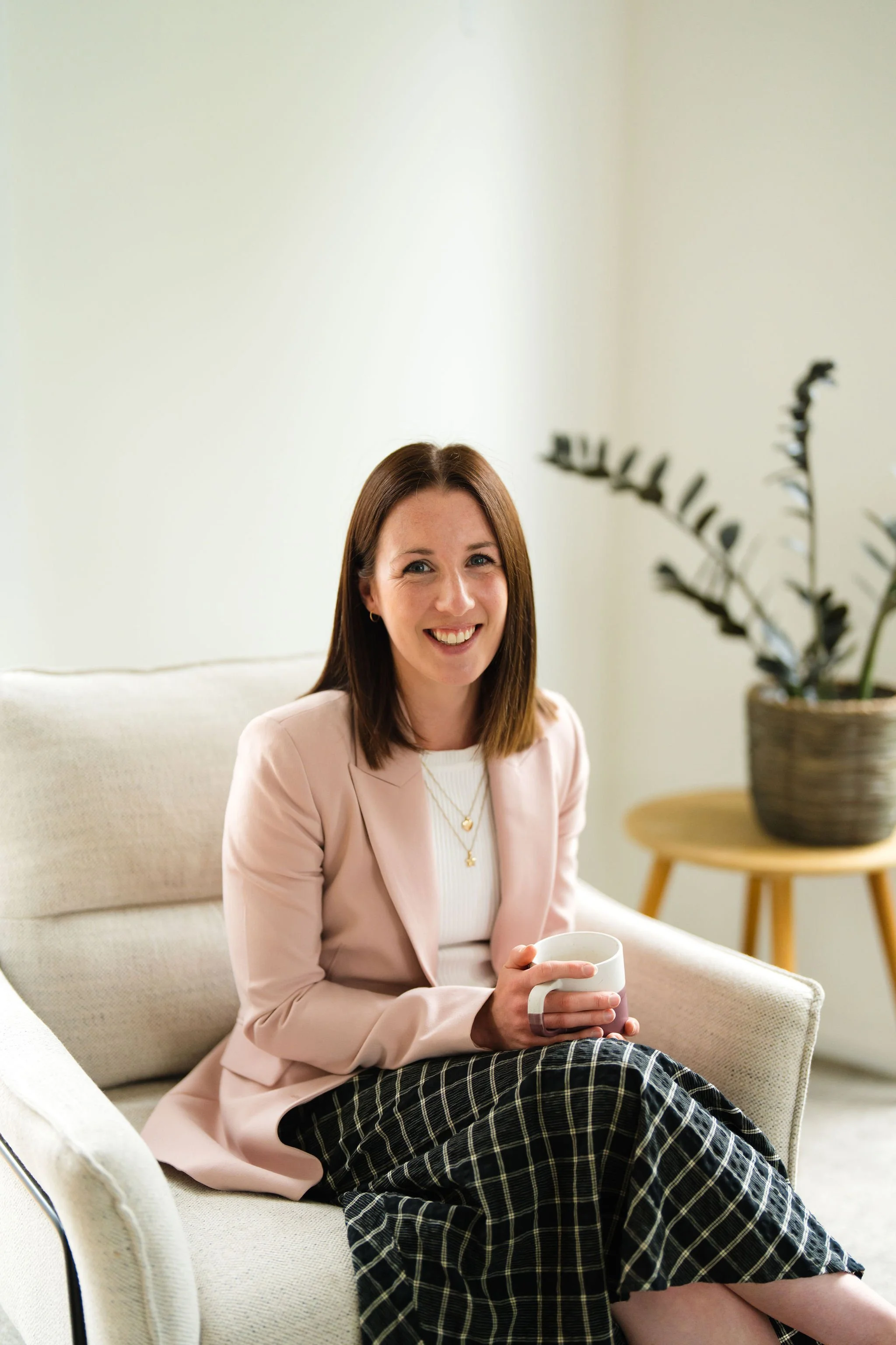 A woman with shoulder-length brown hair, wearing a light pink blazer and black checkered pants, sitting on a cream-colored sofa in a living room, smiling, holding a white mug, with a potted plant on a small wooden table in the background.